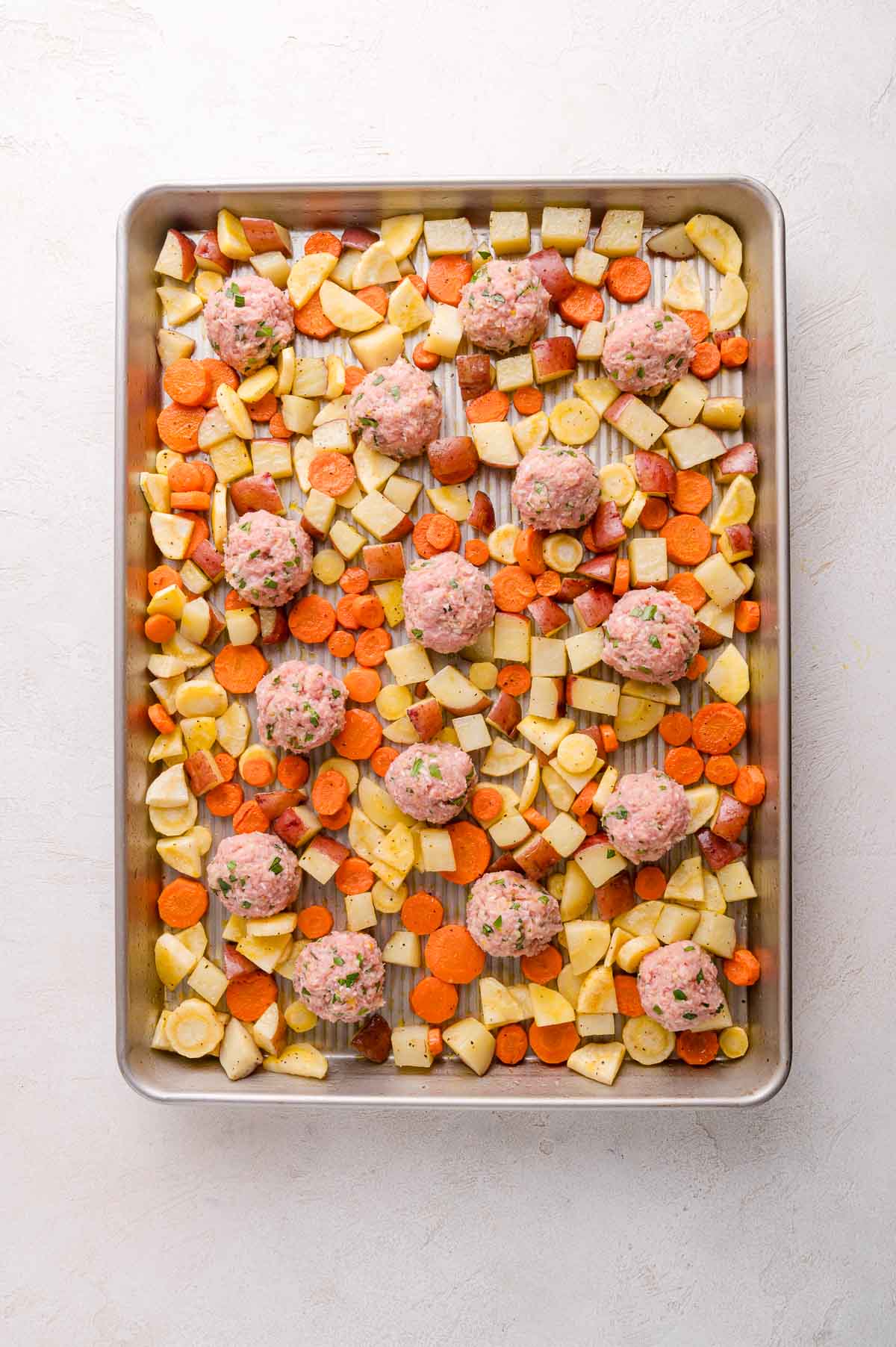 A baking sheet with raw meatballs placed on top of chopped carrots, potatoes, and parsnips, ready to be cooked.