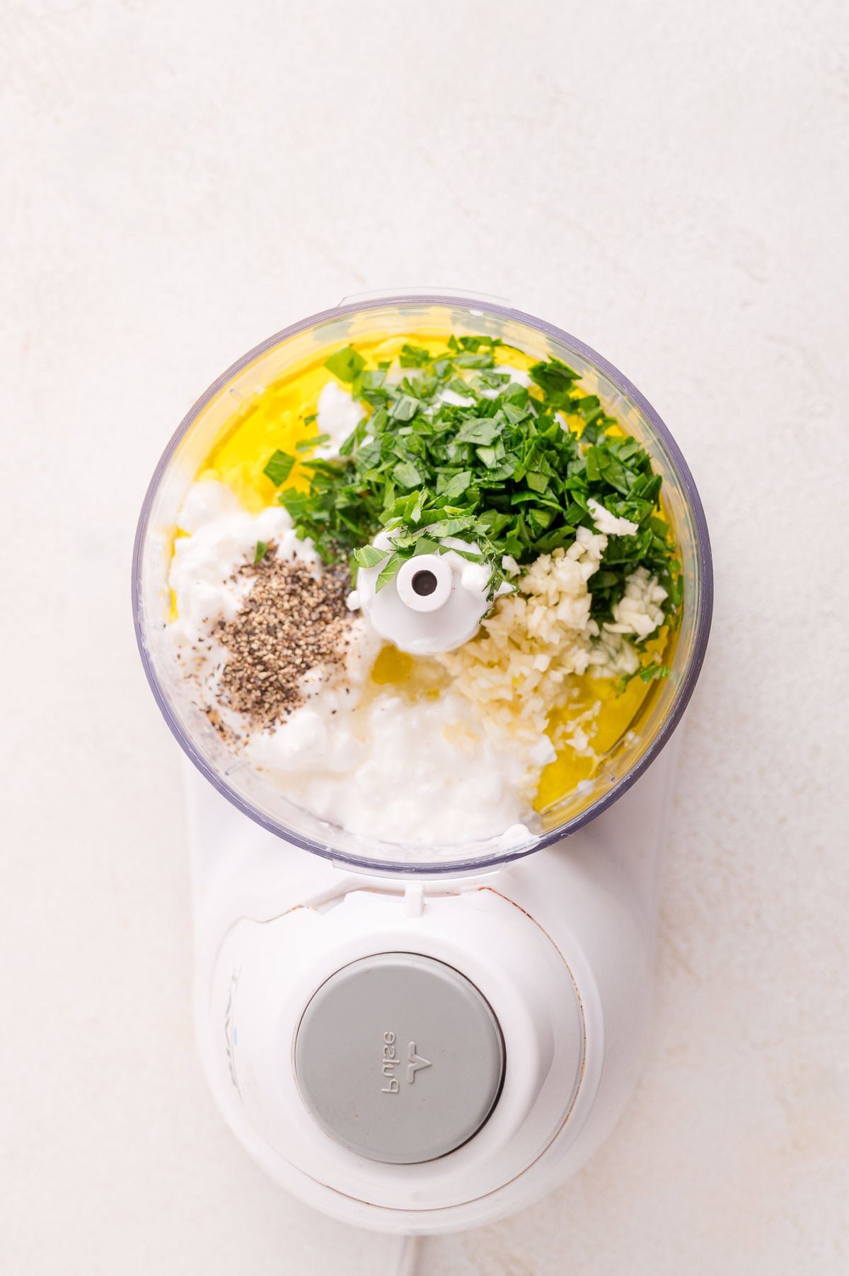A food processor containing cottage cheese, chopped herbs, minced garlic, black pepper, and olive oil, photographed from above.