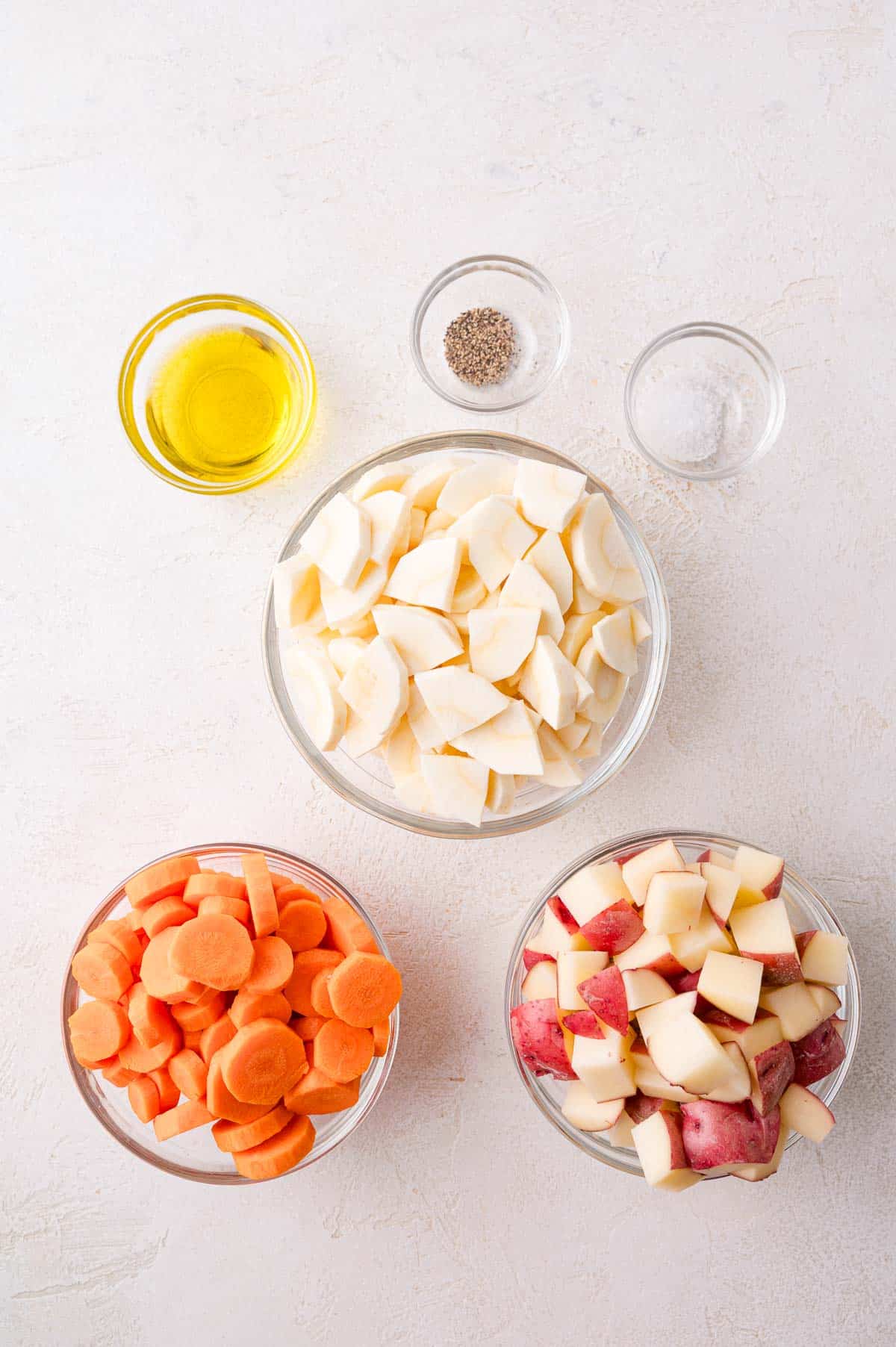Five bowls containing olive oil, ground pepper, salt, chopped parsnips, chopped carrots, and chopped red potatoes arranged on a white surface.