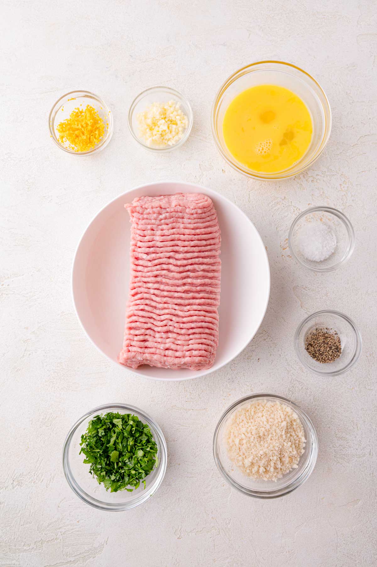 A bowl of ground turkey surrounded by small bowls containing eggs, chopped parsley, breadcrumbs, minced garlic, lemon zest, salt, and pepper on a white surface.