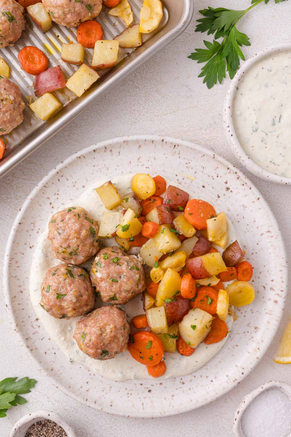 A plate with four meatballs, roasted carrots and potatoes on a cottage cheese sauce, with a tray of more food and bowls of sauce and herbs nearby.
