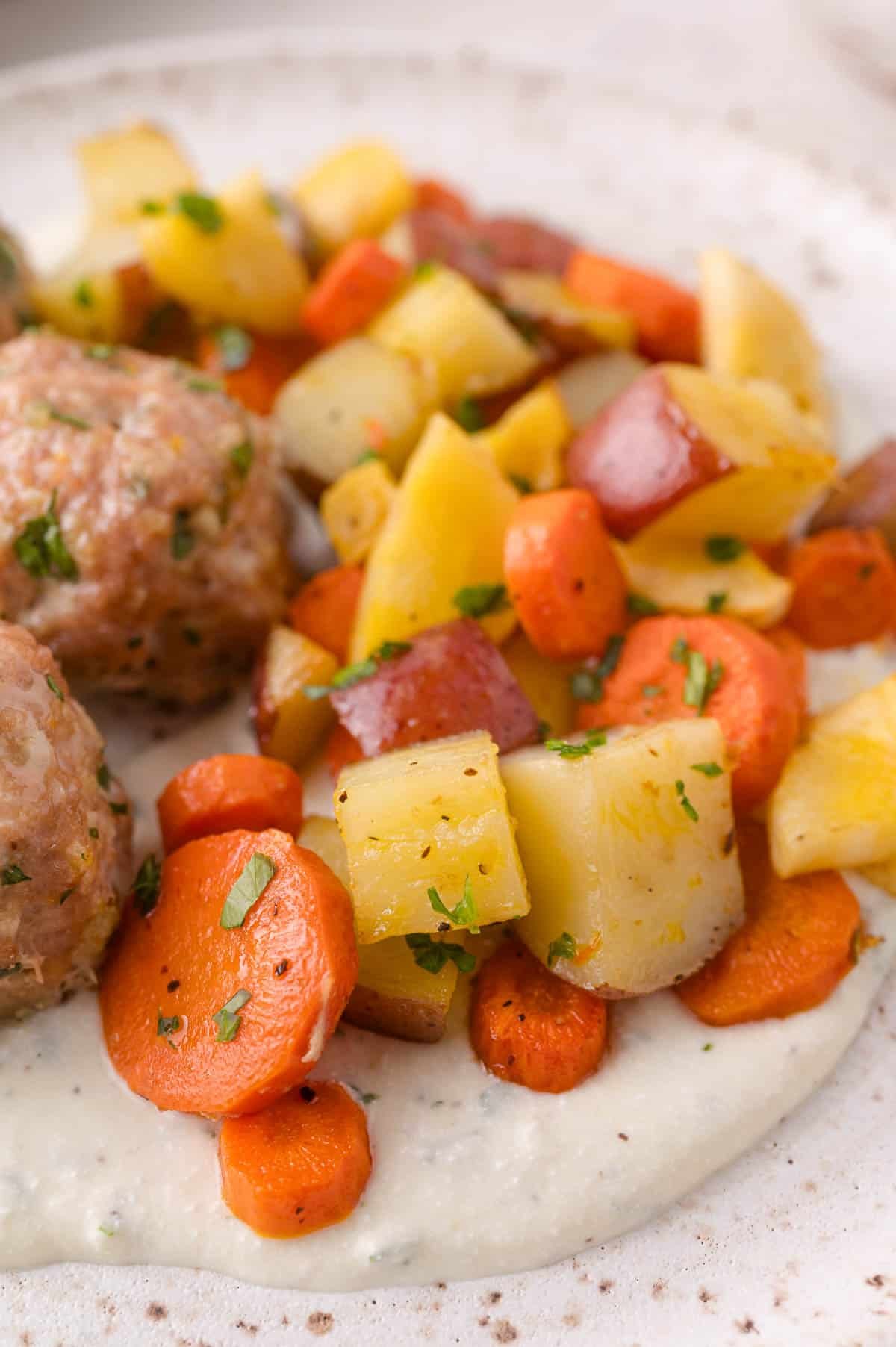 A close-up of roasted carrots, potatoes, and yellow vegetables served over a creamy white sauce, with a meatball partially visible on the side.