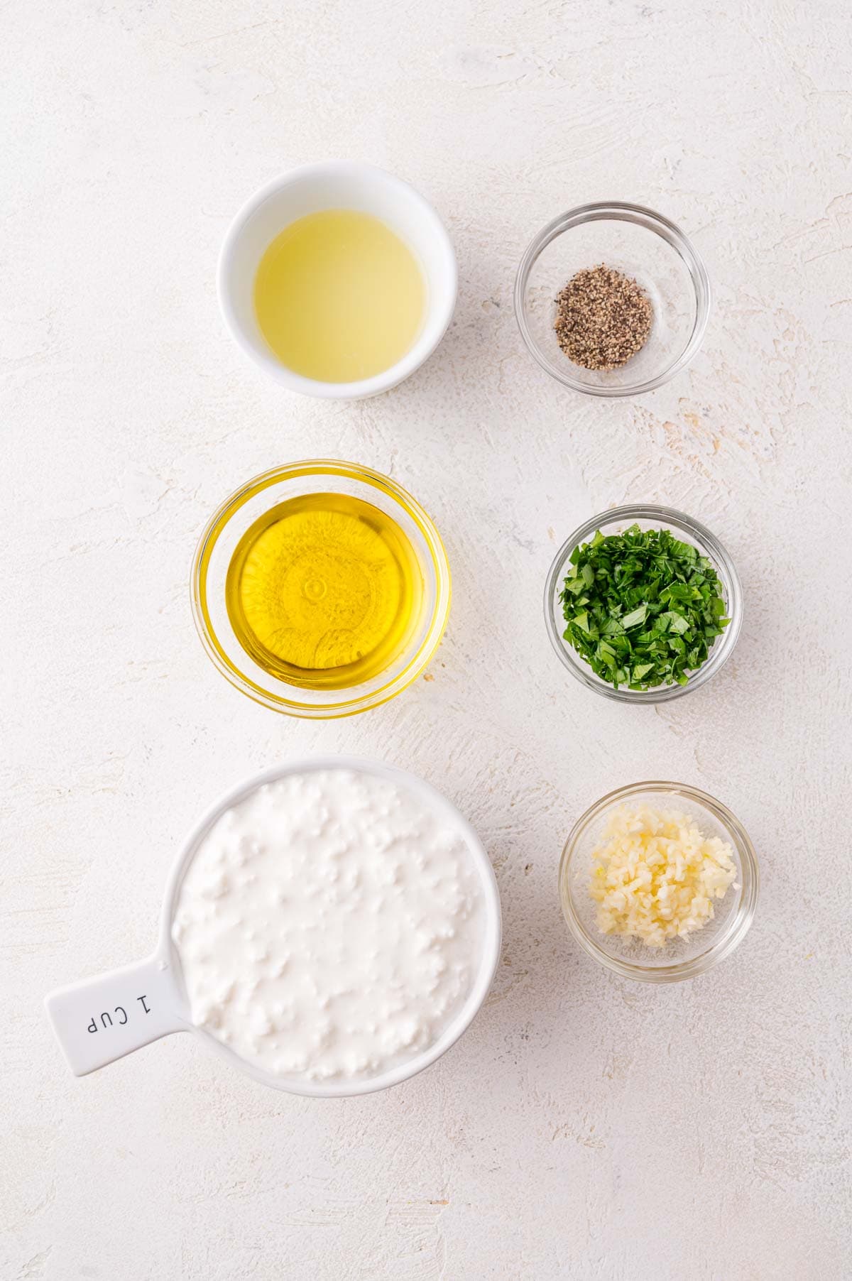Six small bowls contain cottage cheese, olive oil, lemon juice, chopped parsley, minced garlic, and ground black pepper on a light background.