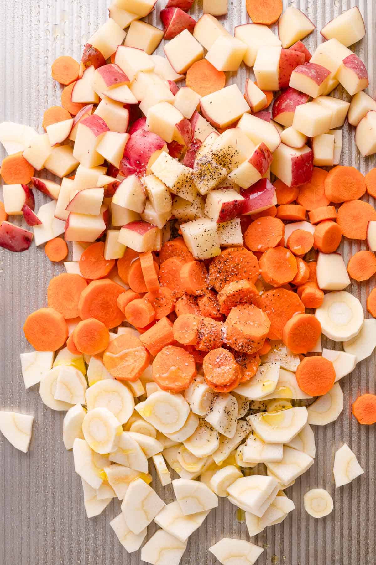 Chopped potatoes, carrots, and parsnips seasoned with ground black pepper on a metal baking sheet.