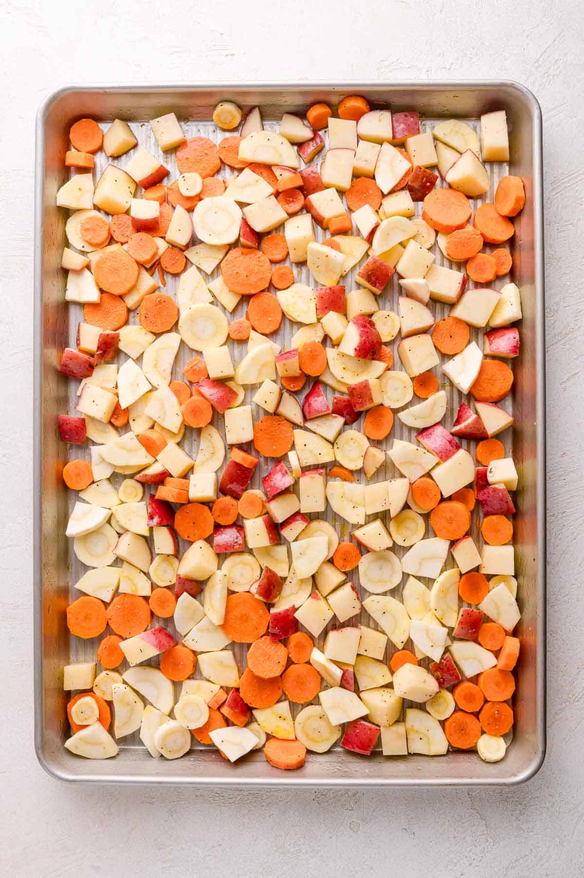 A baking sheet filled with an even layer of chopped root vegetables, including carrots, parsnips, and potatoes, seasoned and ready for roasting.