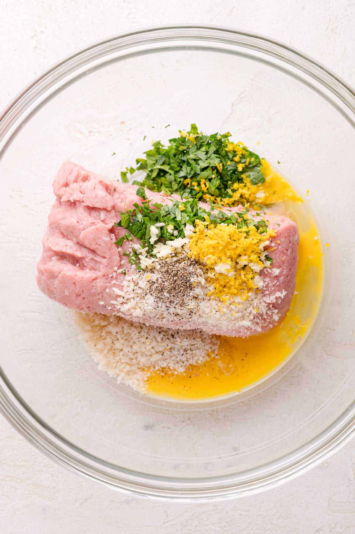 Ground meat, breadcrumbs, chopped herbs, grated lemon zest, black pepper, and a beaten egg in a clear glass mixing bowl on a white surface.