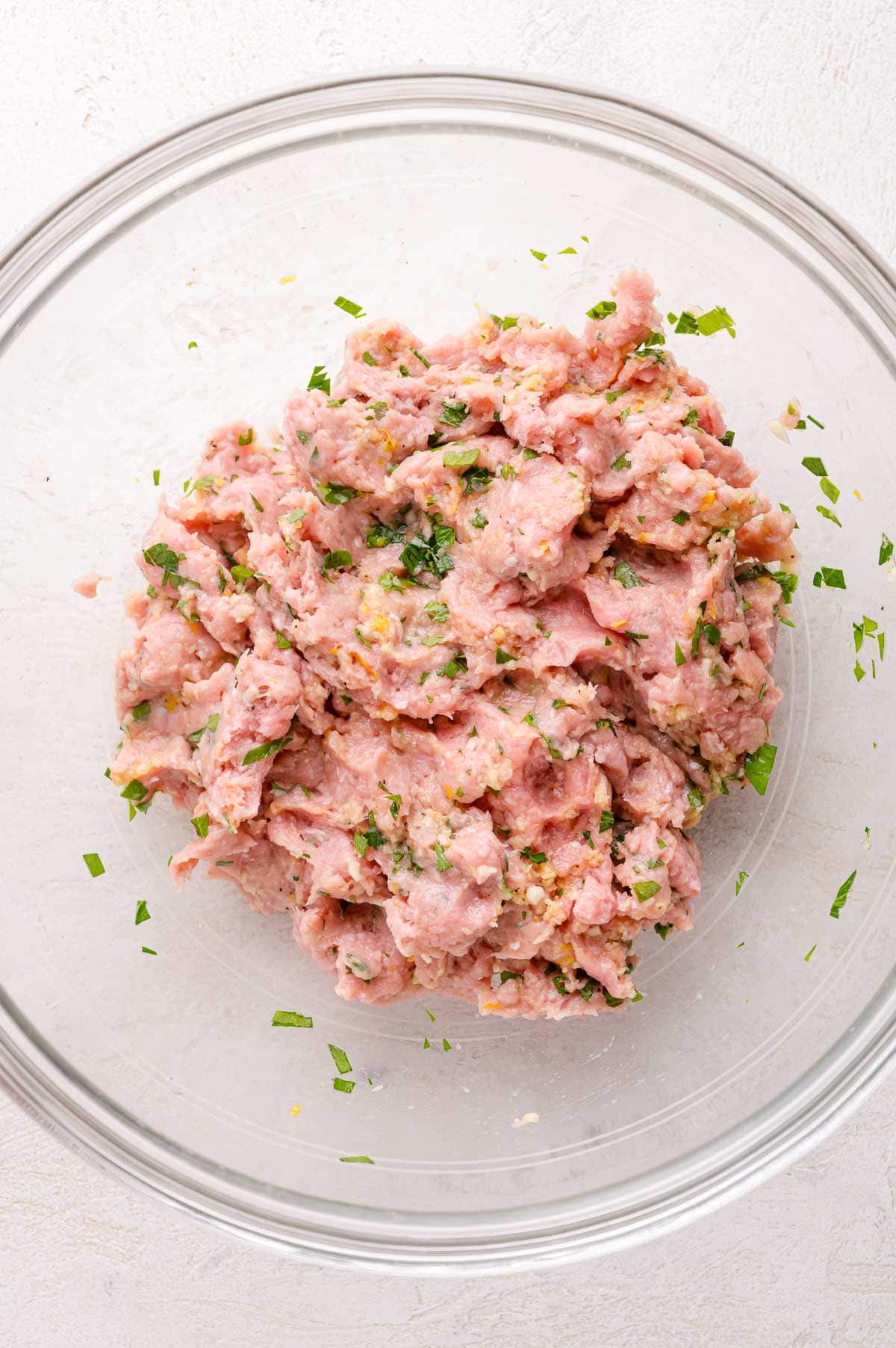A glass bowl filled with seasoned raw ground meat mixed with chopped herbs on a light surface.