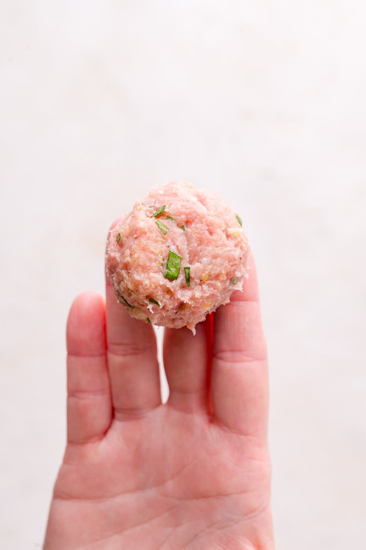 A hand holding a raw meatball mixture with visible green herb pieces against a light background.