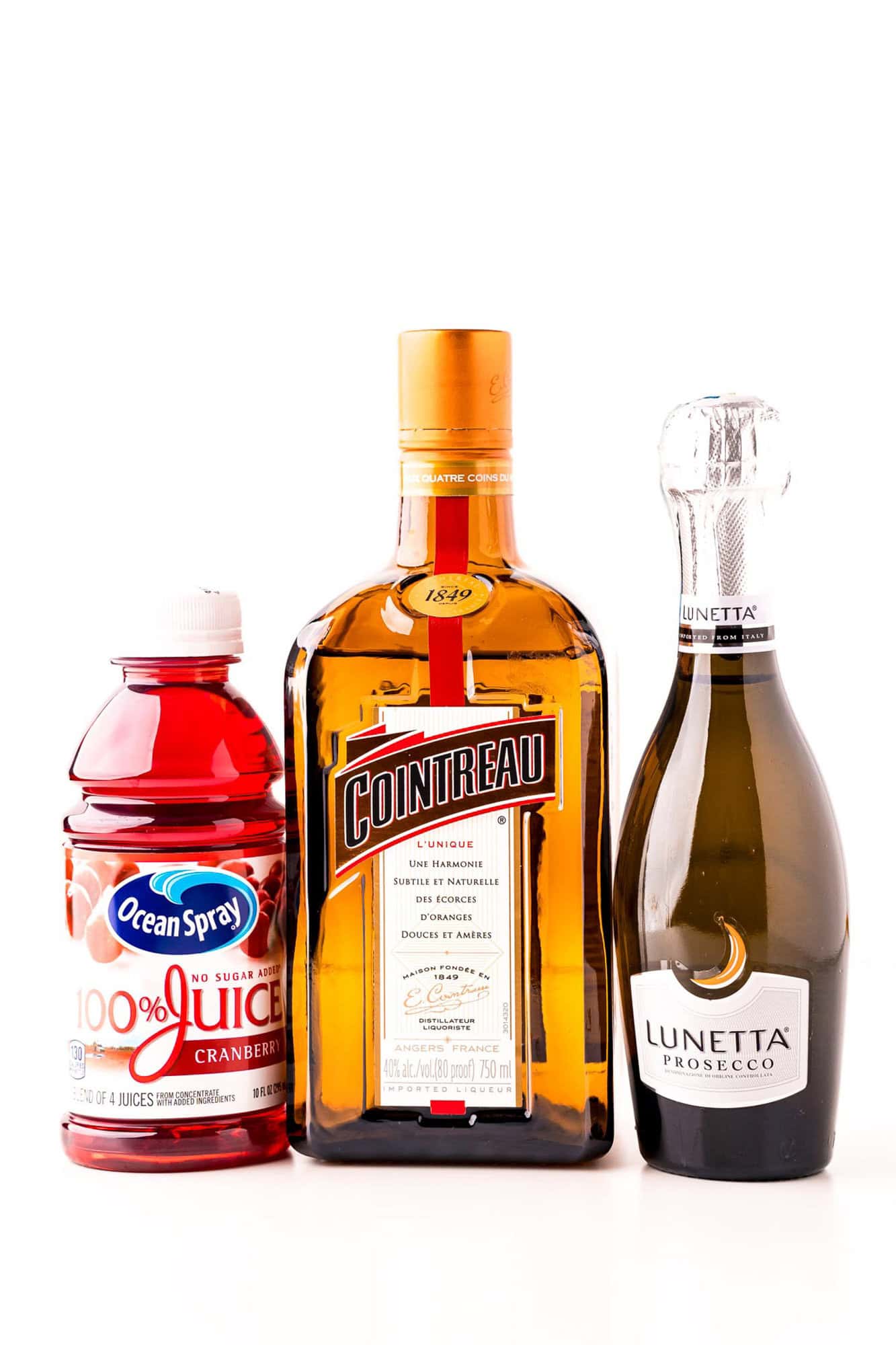 A bottle of cranberry juice, a bottle of Cointreau liqueur, and a bottle of Lunetta Prosecco are shown side by side against a white background.