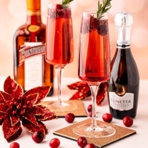Two champagne flutes filled with a red poinsettia cocktail garnished with cranberries and rosemary, next to bottles of Cointreau and Prosecco, with festive red decorations and loose cranberries.