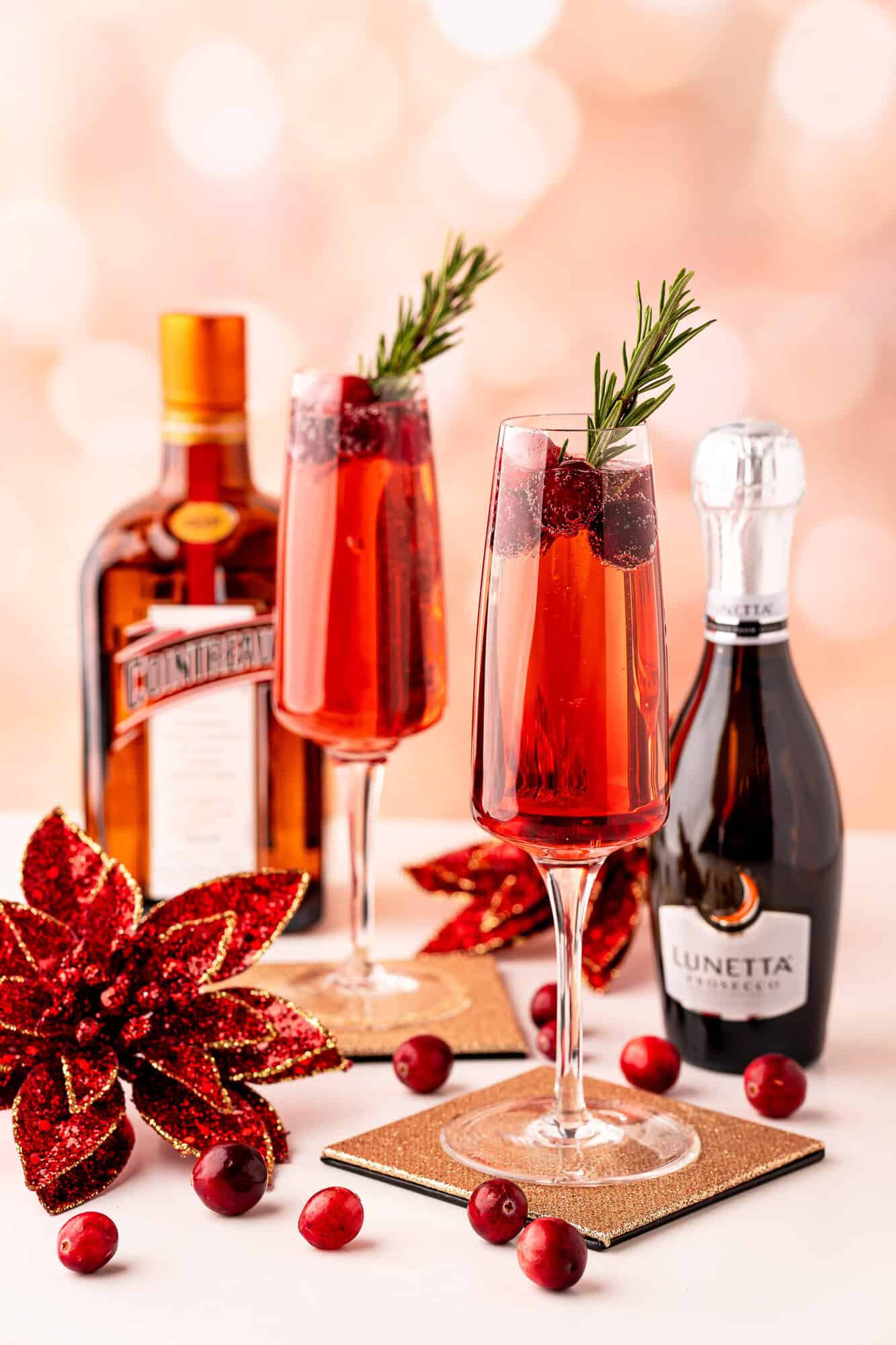 Two glasses of red cocktails with cranberries and rosemary garnish next to Cointreau and Lunetta bottles, decorative poinsettia, and scattered cranberries.