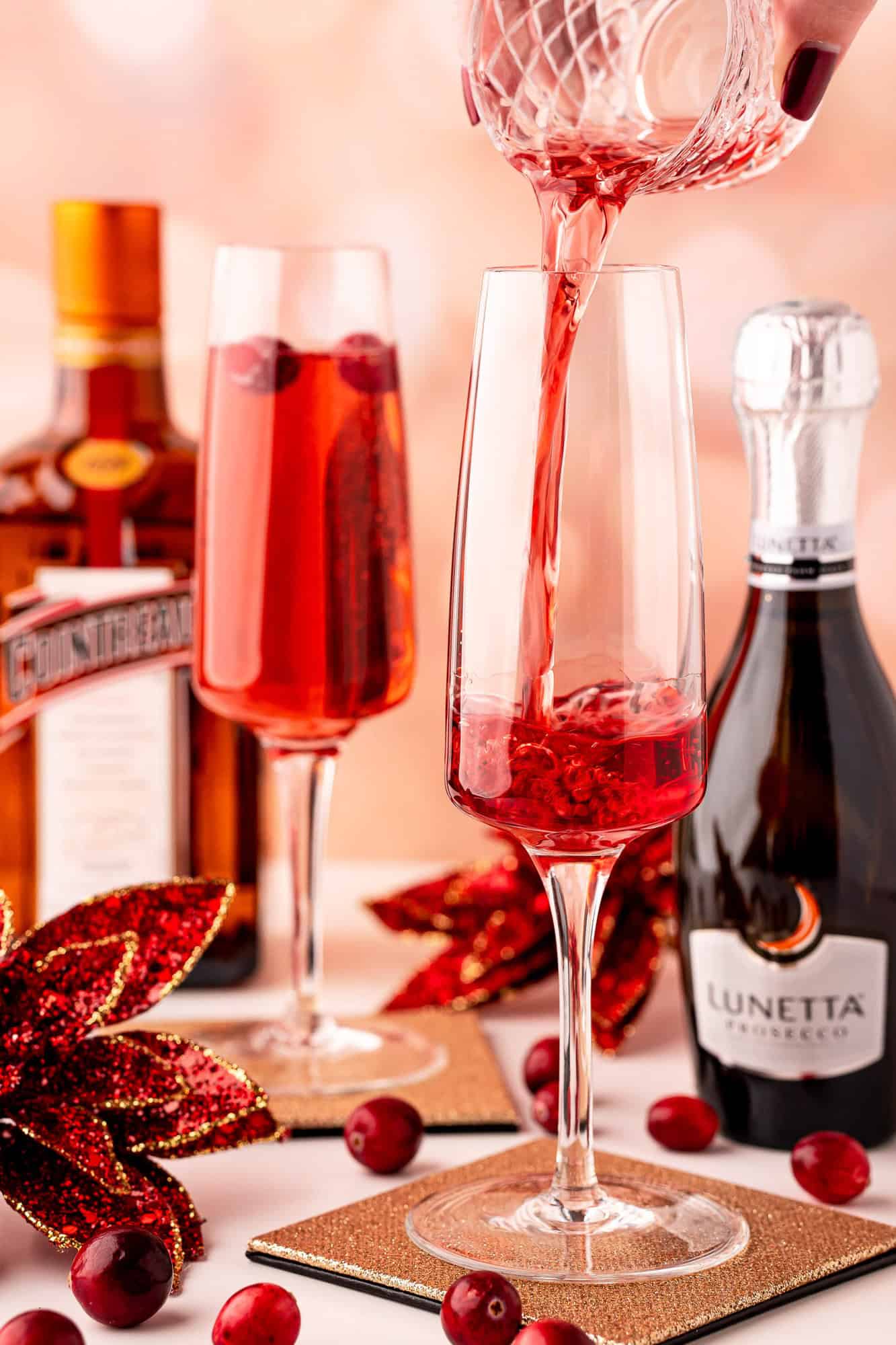 Cranberry juice being poured into a champagne flute, with another filled glass, Cointreau and Lunetta Prosecco bottles, cranberries, and holiday decorations on the table.