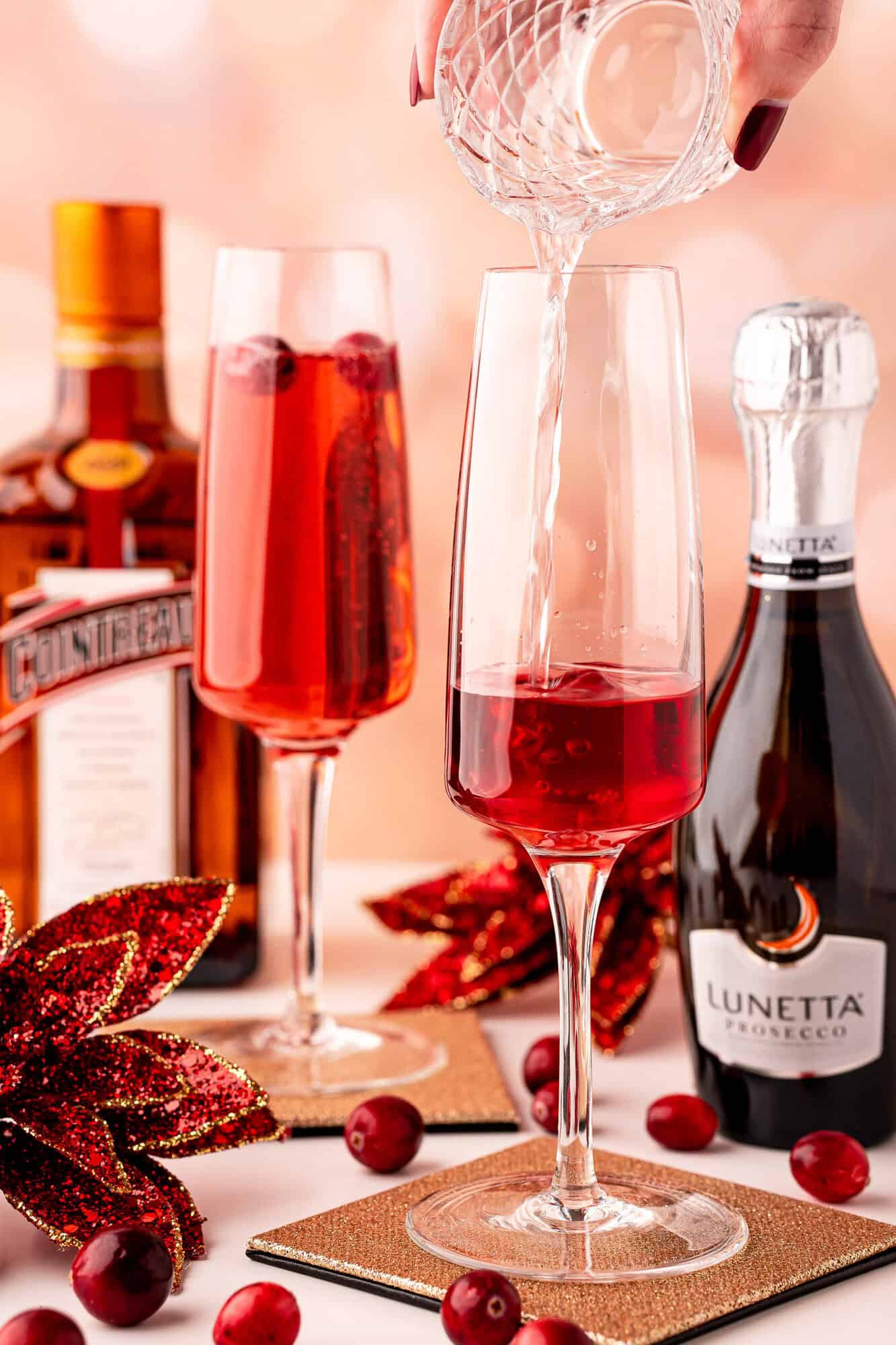 A hand pours Cointreau into a flute glass with red drink, next to another filled flute, bottles of Cointreau and Prosecco, cranberries, and festive decorations.