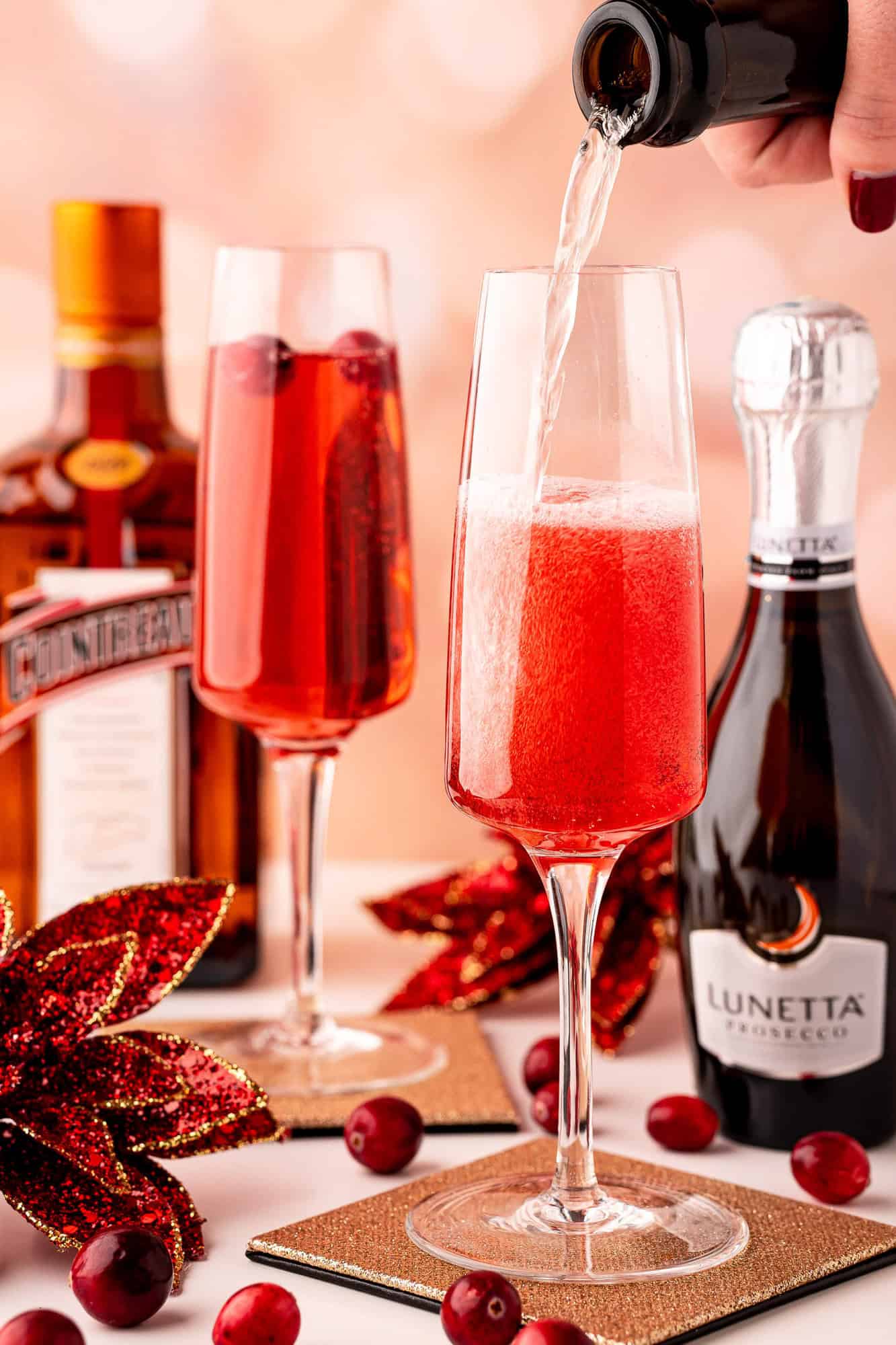 A hand pours sparkling wine into a glass with red liquid and cranberries. A bottle of prosecco, Cointreau, and festive decorations are on the table.