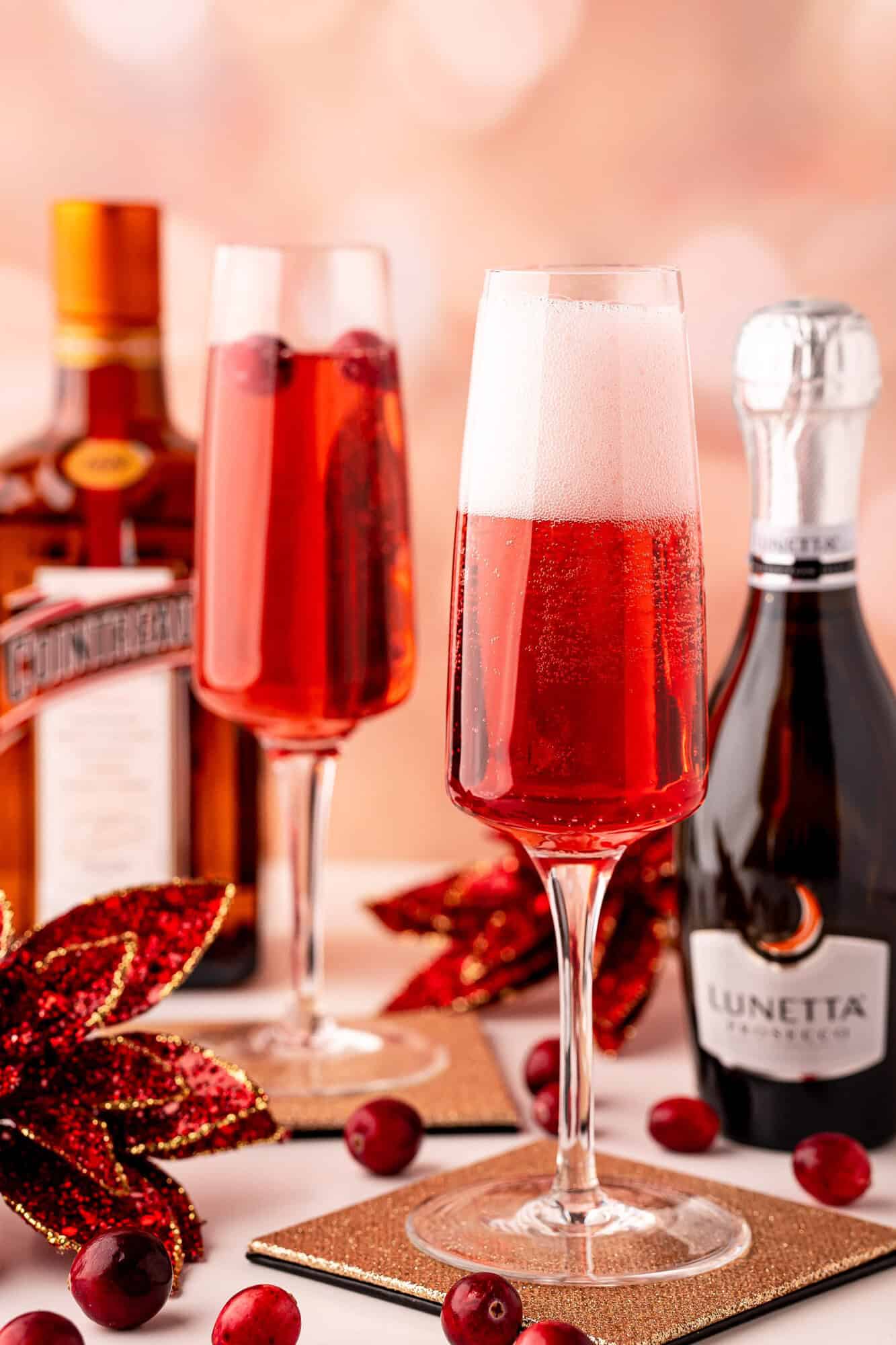 Two champagne flutes with red sparkling cocktails, garnished with cranberries, sit beside bottles of Cointreau and Lunetta Prosecco, with festive decorations in the background.