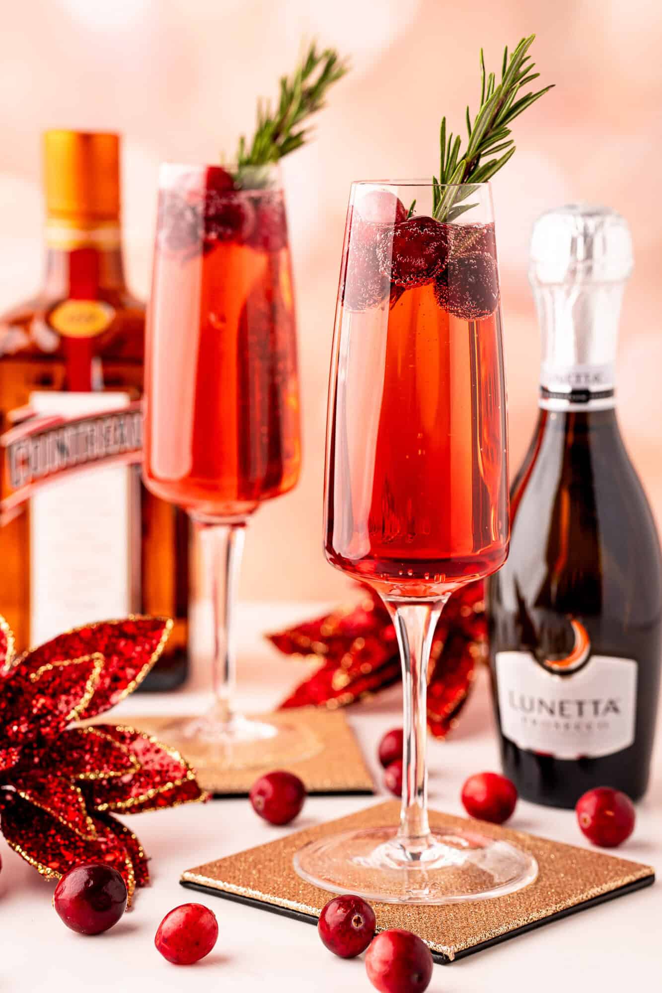 Two champagne flutes with red poinsettia cocktails, garnished with cranberries and rosemary, are placed on coasters beside a Cointreau bottle, a Lunetta Prosecco bottle, and decorative poinsettias.