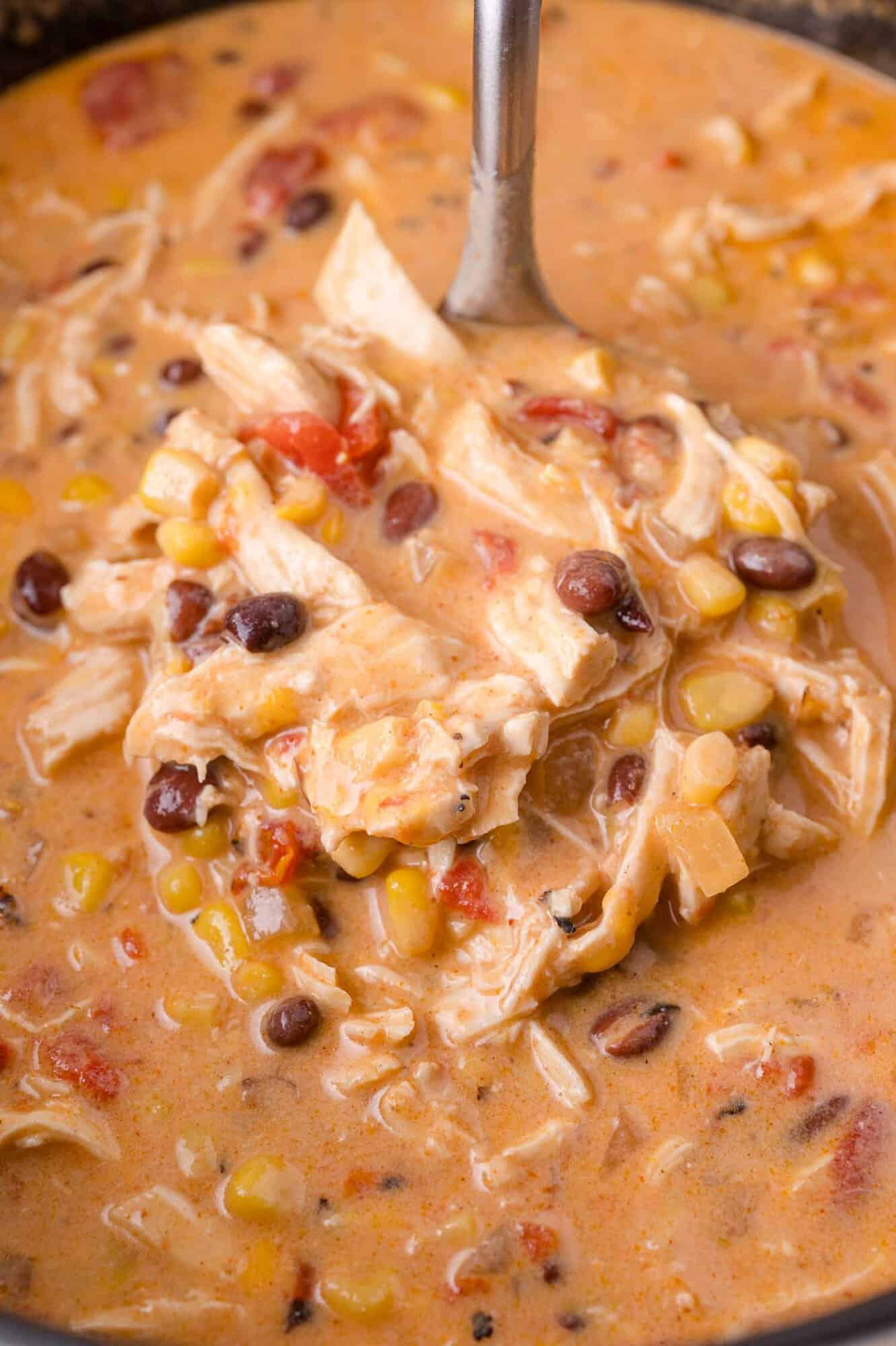 A ladle scoops creamy soup with shredded chicken, black beans, corn, and diced tomatoes from a pot.