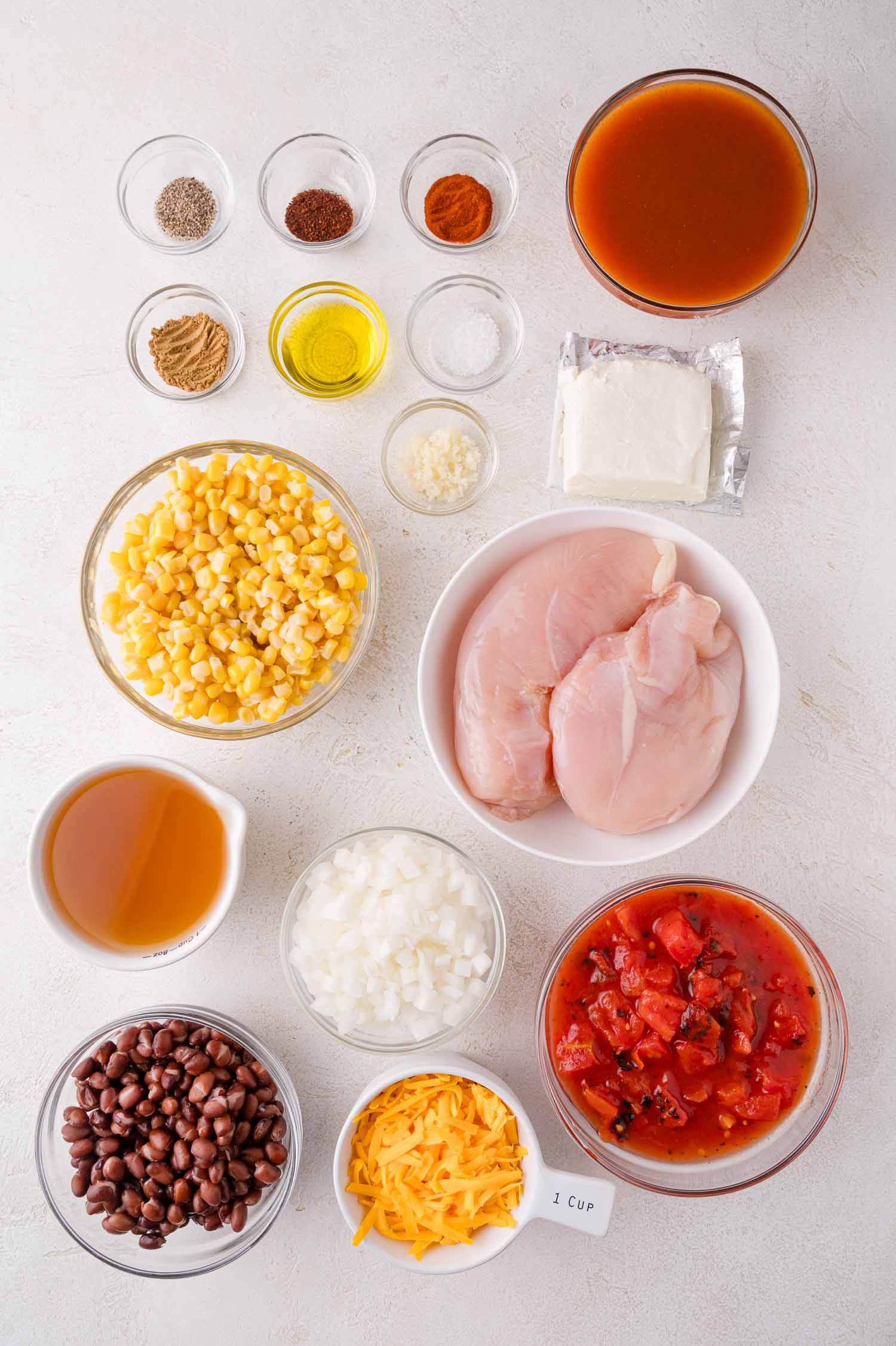 Various ingredients for chicken enchilada soup recipe are arranged on a white surface, including raw chicken breasts, black beans, corn, diced tomatoes, cheese, broth, seasonings, and chopped onion.