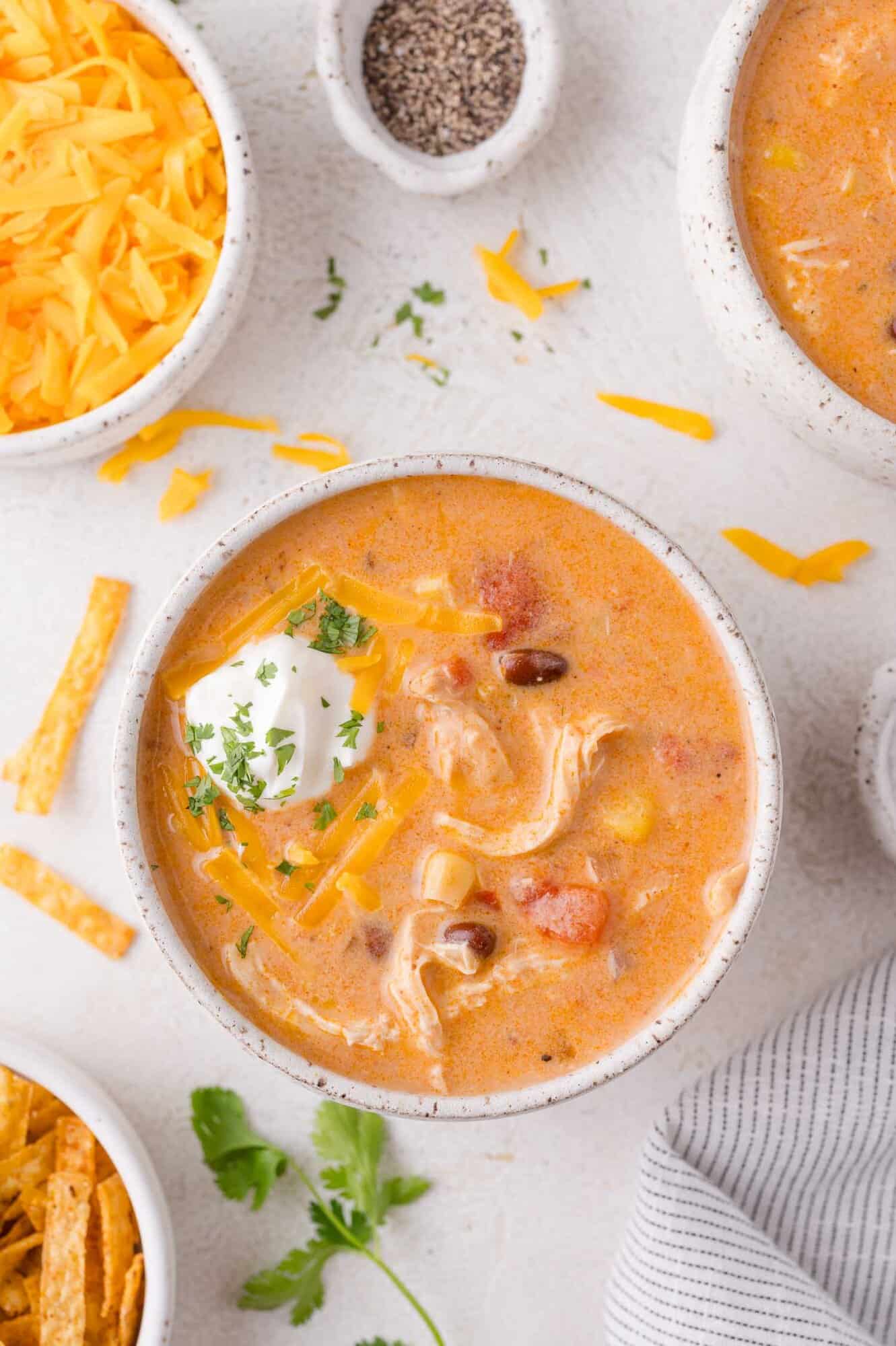 Bowl of creamy chicken enchilada soup with shredded chicken, beans, corn, tomatoes, shredded cheese, and a dollop of sour cream, surrounded by bowls of grated cheese, tortilla strips, and pepper.