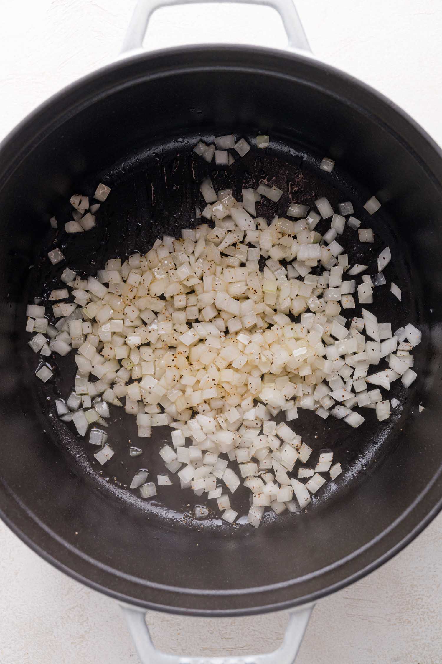 Chopped onions are being sautéed with black pepper in a large black pot on a light-colored surface.