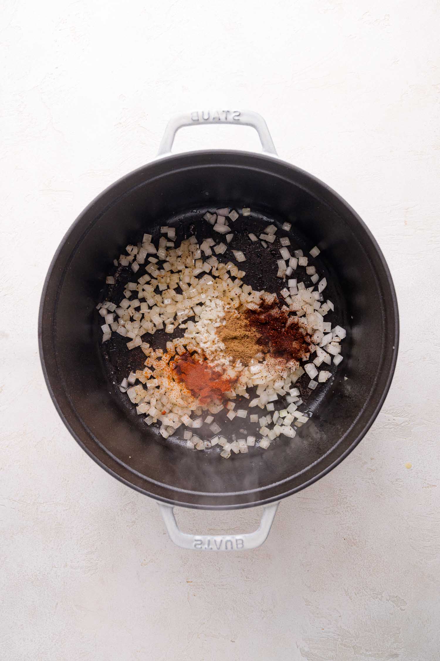 Diced onions and assorted spices are cooking in a black pot on a light-colored surface.