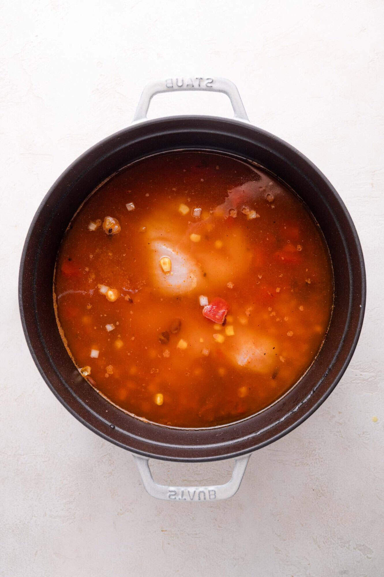 A black pot filled with a tomato-based soup containing corn, beans, tomatoes, and chunks of chicken, viewed from above.