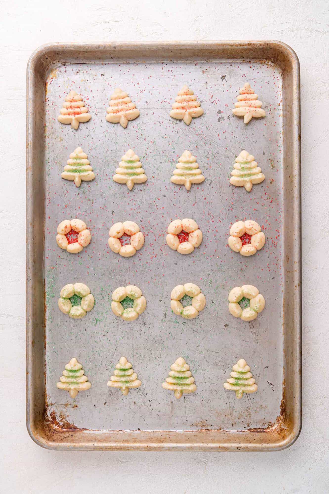 A baking sheet with rows of small Christmas tree and wreath-shaped cookies, lightly decorated with red and green sugar.