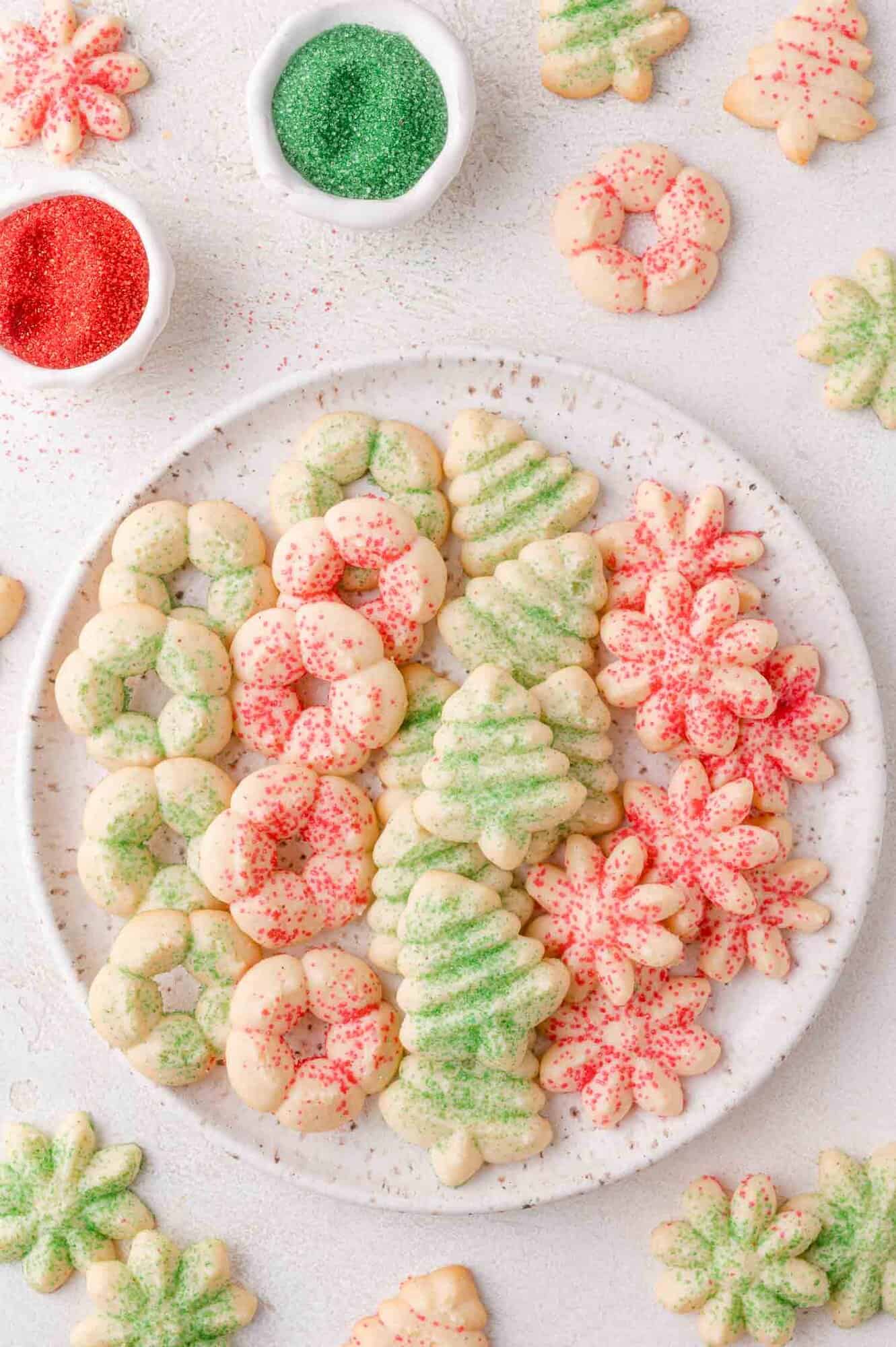 A plate of assorted holiday cookies shaped like trees, flowers, and rings, decorated with red and green sugar sprinkles. Small bowls of colored sugar are nearby.
