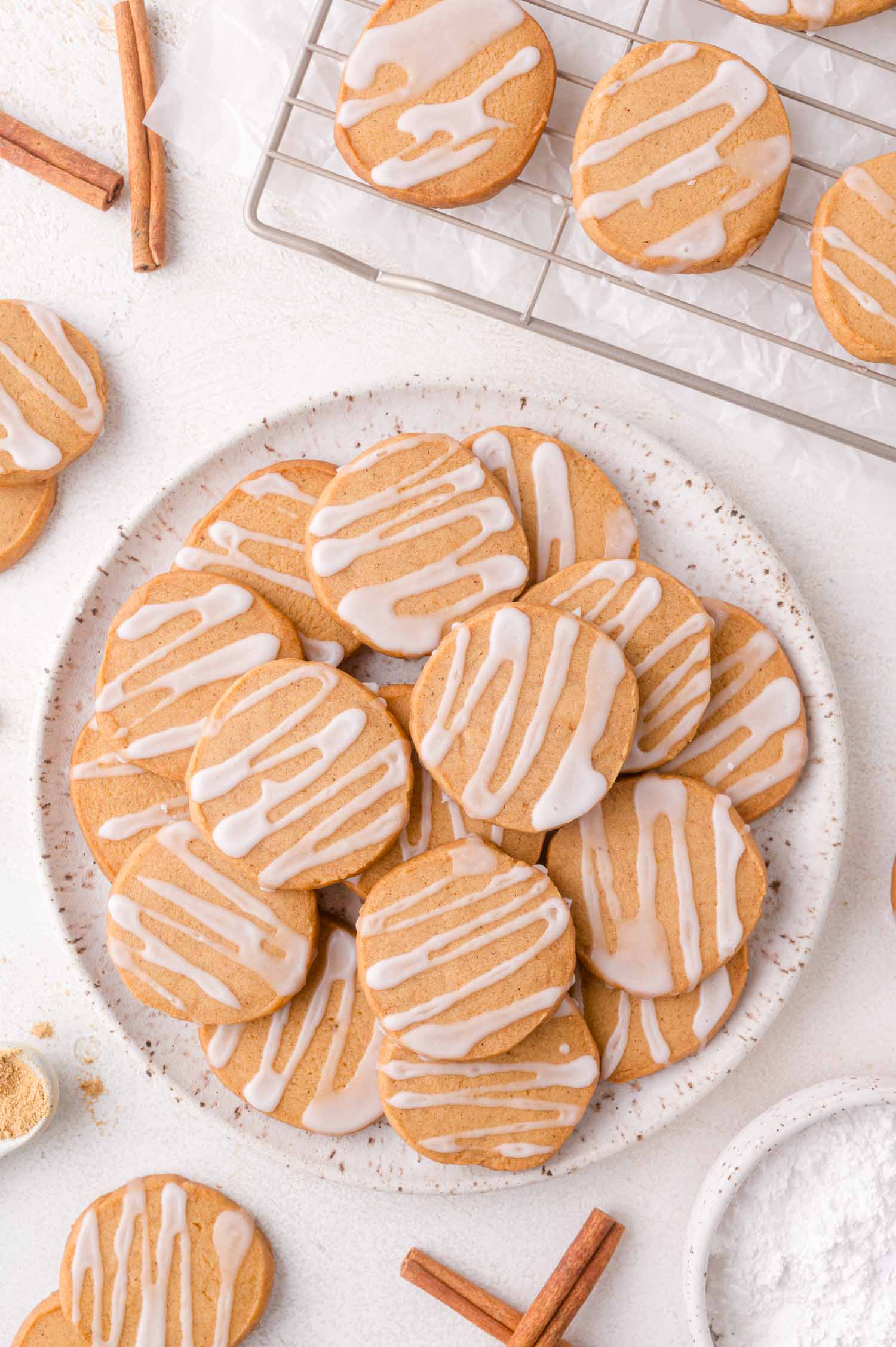 A plate of round gingerbread shortbread cookies with white icing drizzle, surrounded by more cookies, cinnamon sticks, and a cooling rack on a white surface.