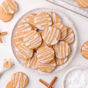 Plate of round gingerbread shortbread cookies with white icing drizzle, surrounded by cinnamon sticks, a cooling rack, and baking ingredients on a white surface.