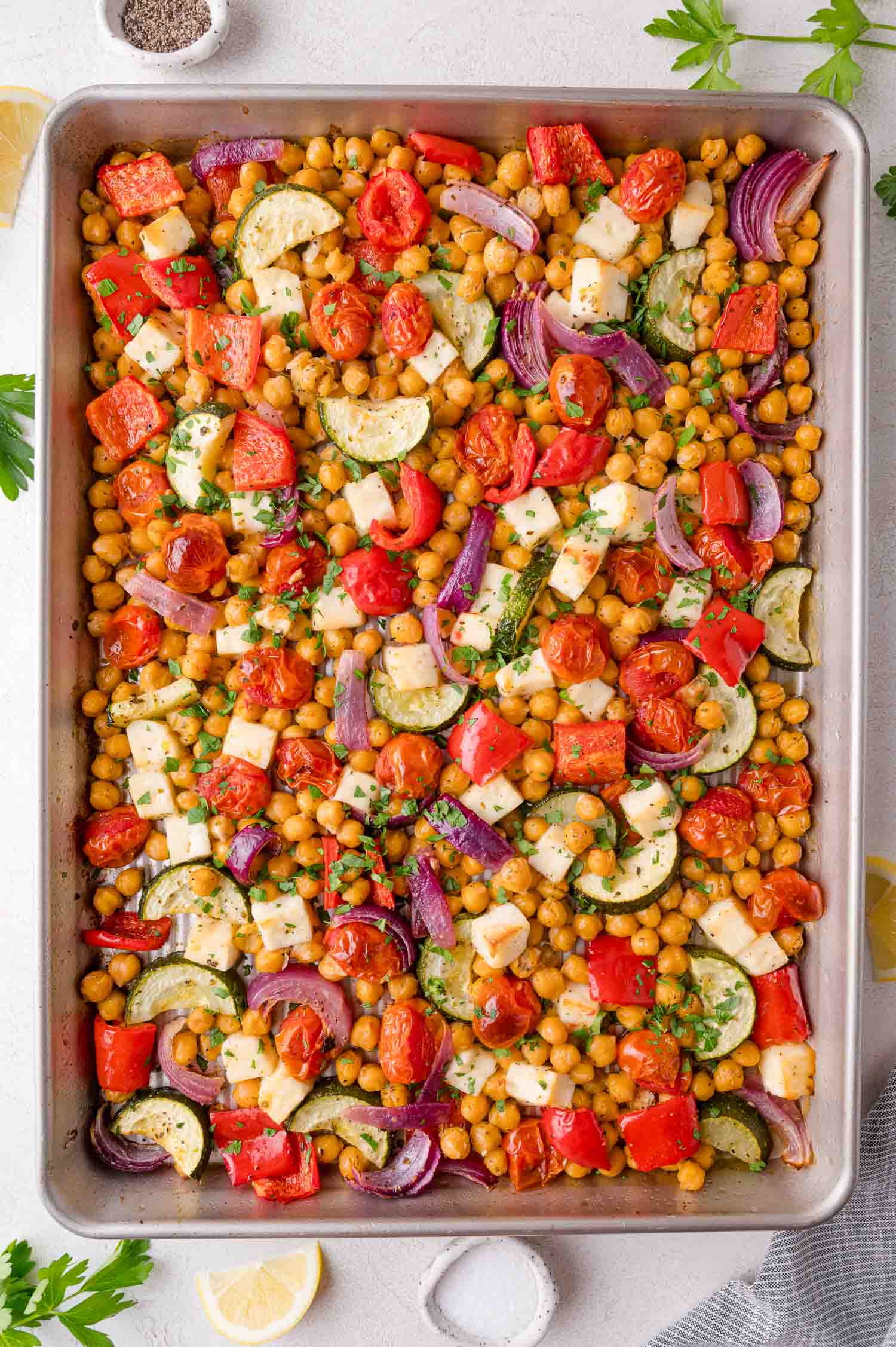 A baking sheet filled with roasted chickpeas, cherry tomatoes, zucchini, red bell peppers, red onions, and cubes of cheese, garnished with chopped parsley.