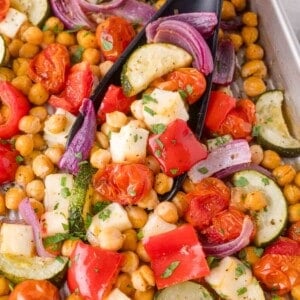 A baking tray with roasted chickpeas, red bell peppers, cherry tomatoes, zucchini, red onion, and cubes of halloumi cheese, garnished with chopped herbs.