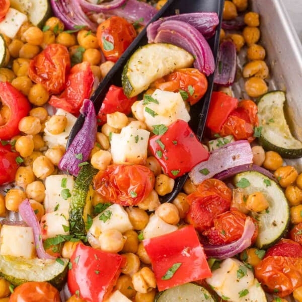 A baking tray with roasted chickpeas, red bell peppers, cherry tomatoes, zucchini, red onion, and cubes of halloumi cheese, garnished with chopped herbs.