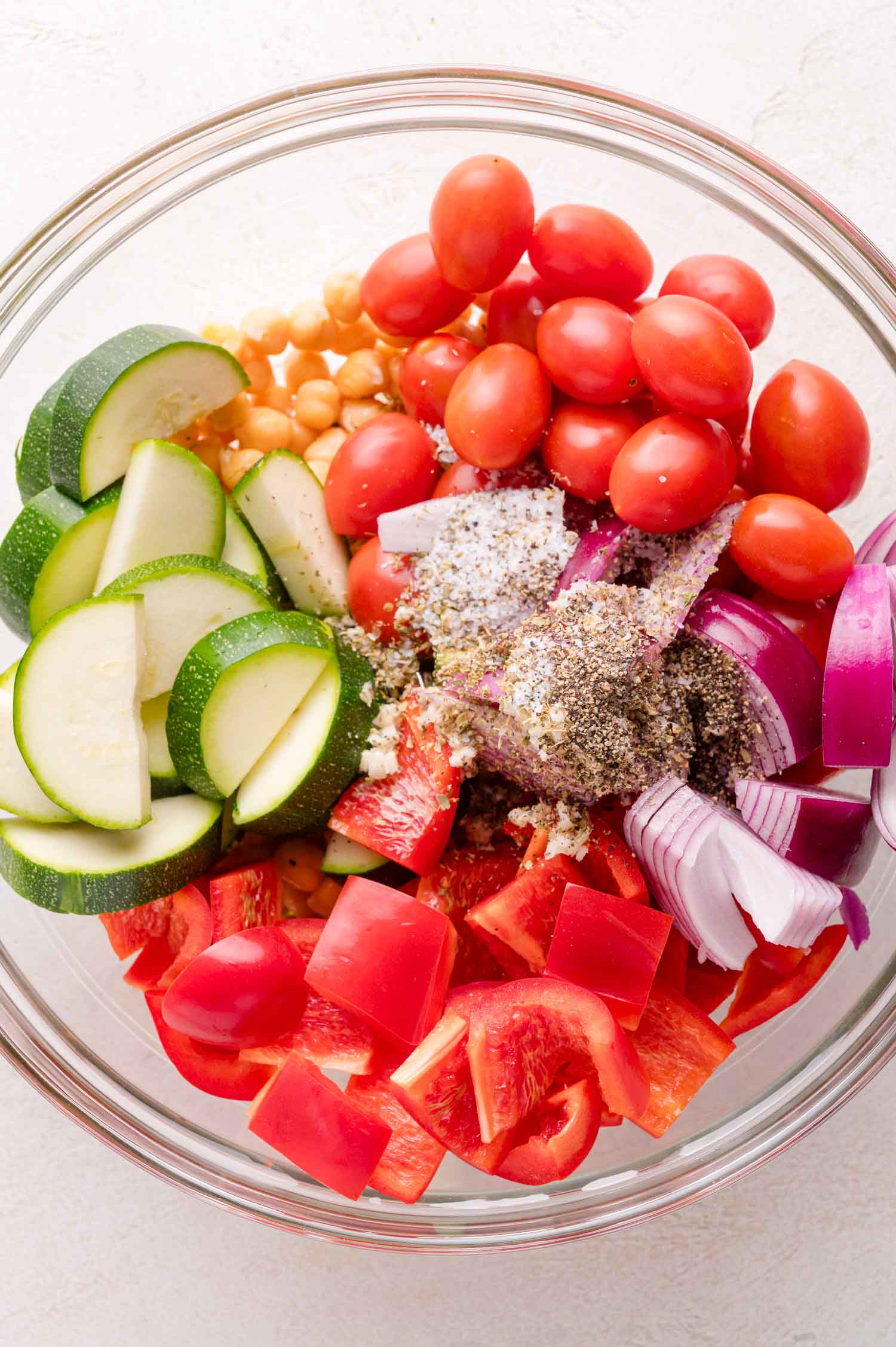 A glass bowl containing grape tomatoes, chickpeas, sliced zucchini, chopped red onion, chopped red bell pepper, and assorted seasonings.