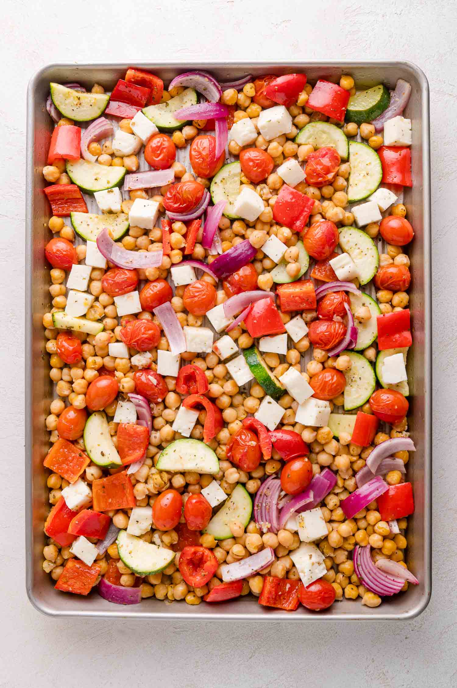 A baking sheet filled with chopped vegetables, cherry tomatoes, red onion, zucchini, bell pepper, chickpeas, and cubes of cheese, all arranged in an even layer.