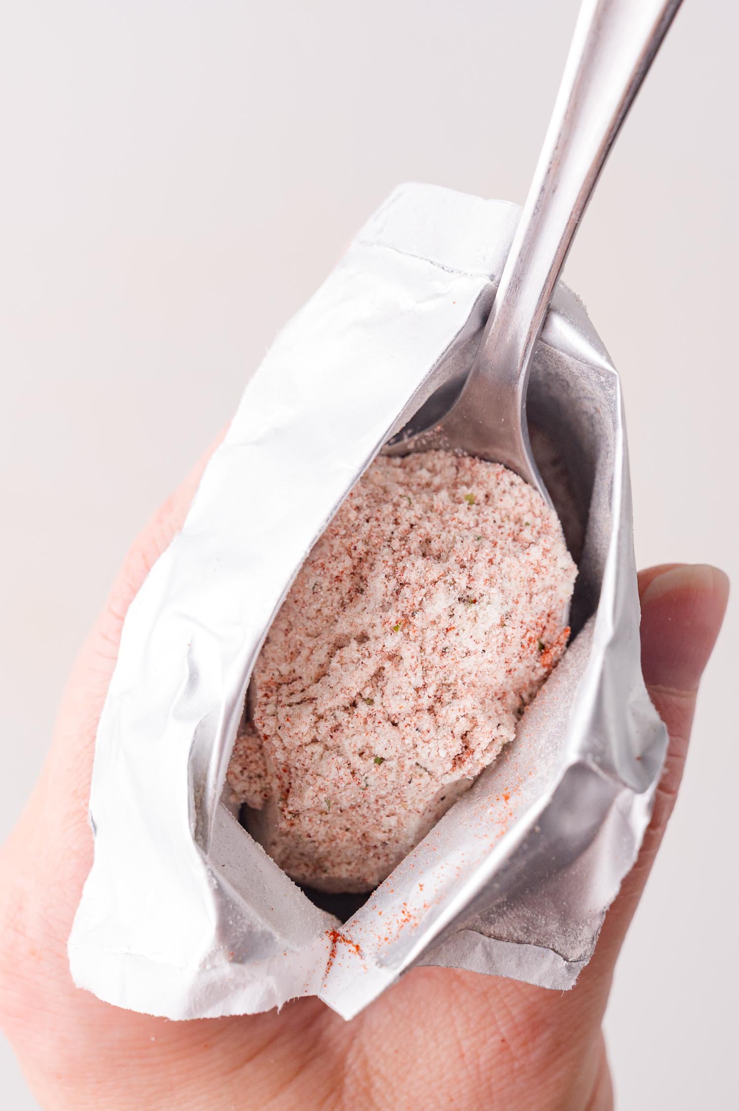 A hand holding an open foil packet of ranch seasoning, with a spoon inside.