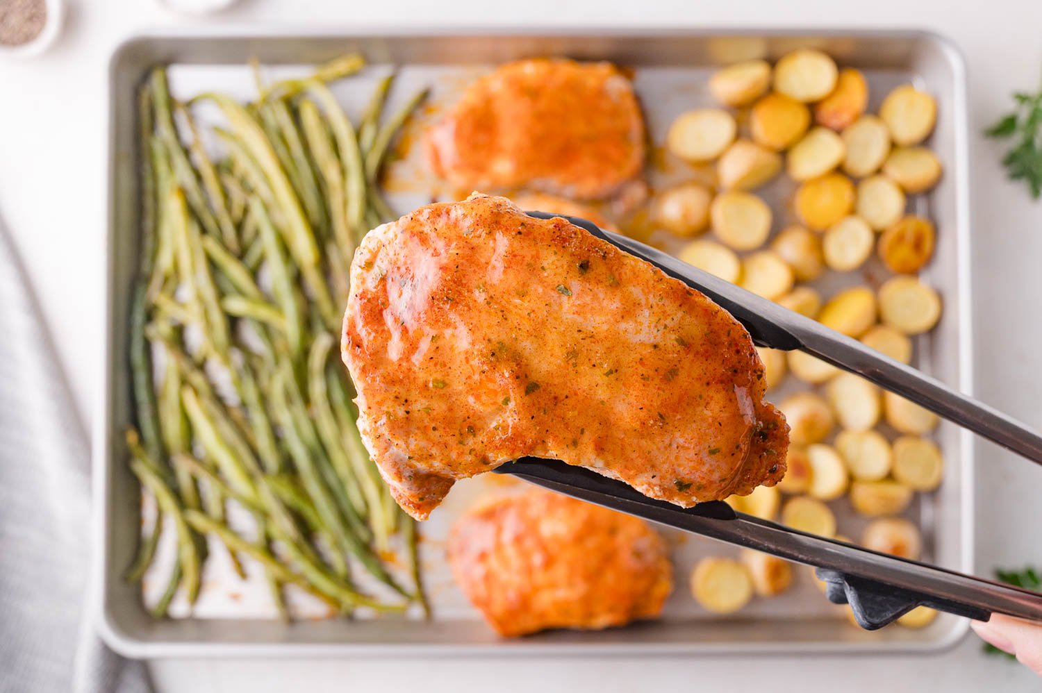 A pair of tongs holds a cooked pork chop above a sheet pan with roasted green beans and sliced potatoes.