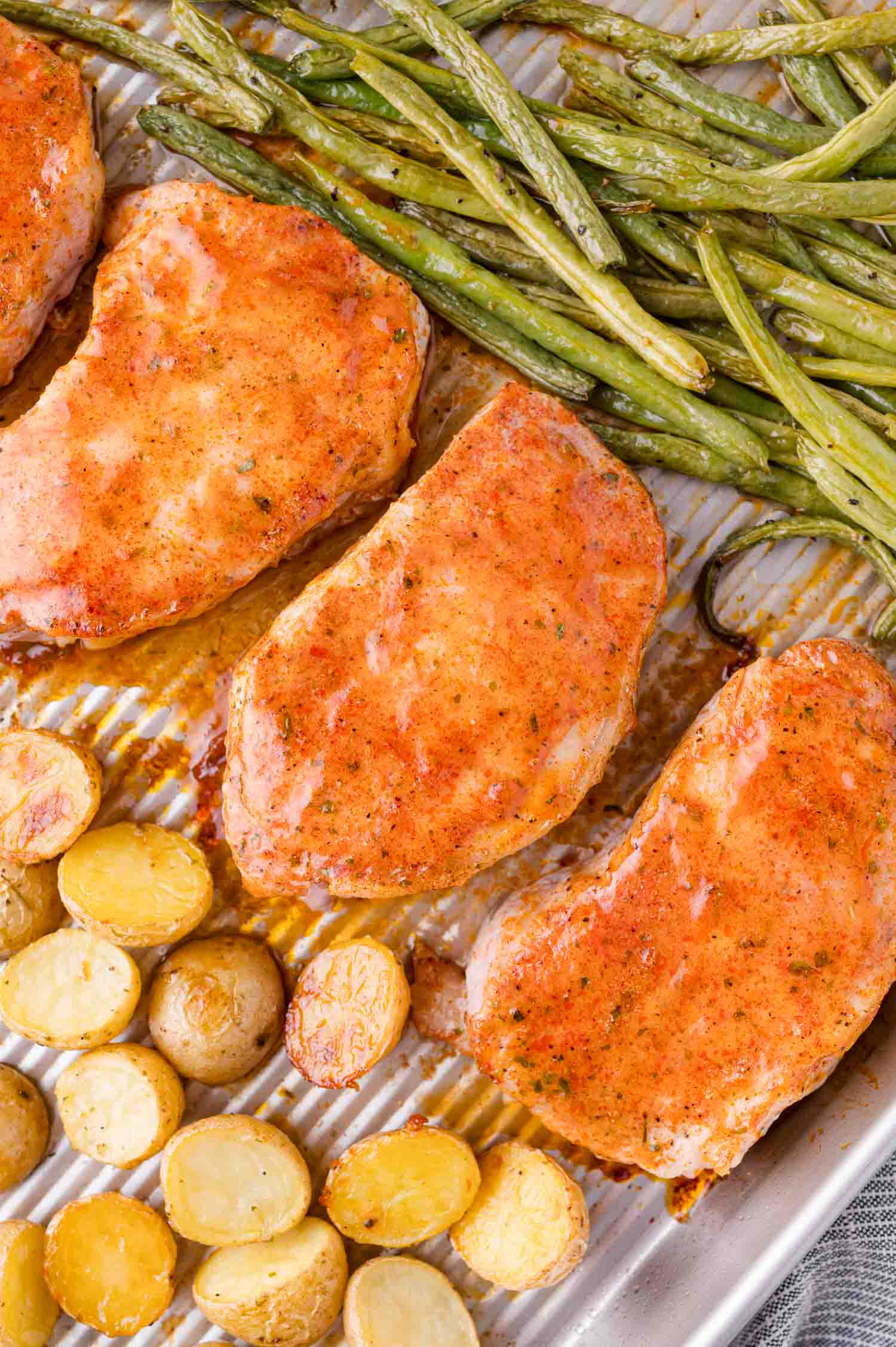 Baked pork chops, green beans, and sliced potatoes arranged on a metal baking sheet.