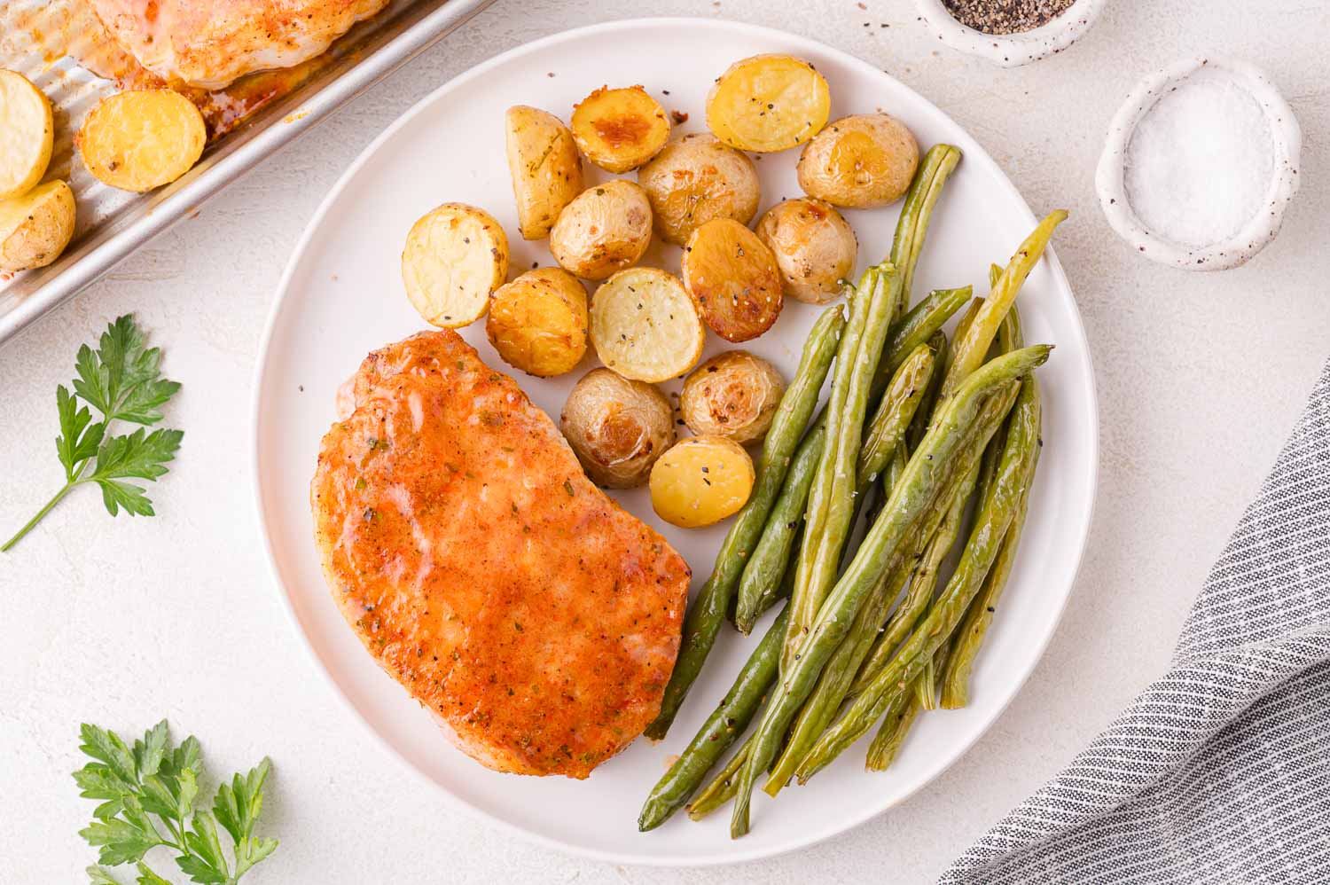 A plate with a baked pork chop, roasted baby potatoes, and green beans, placed next to salt, pepper, parsley, and a striped napkin.
