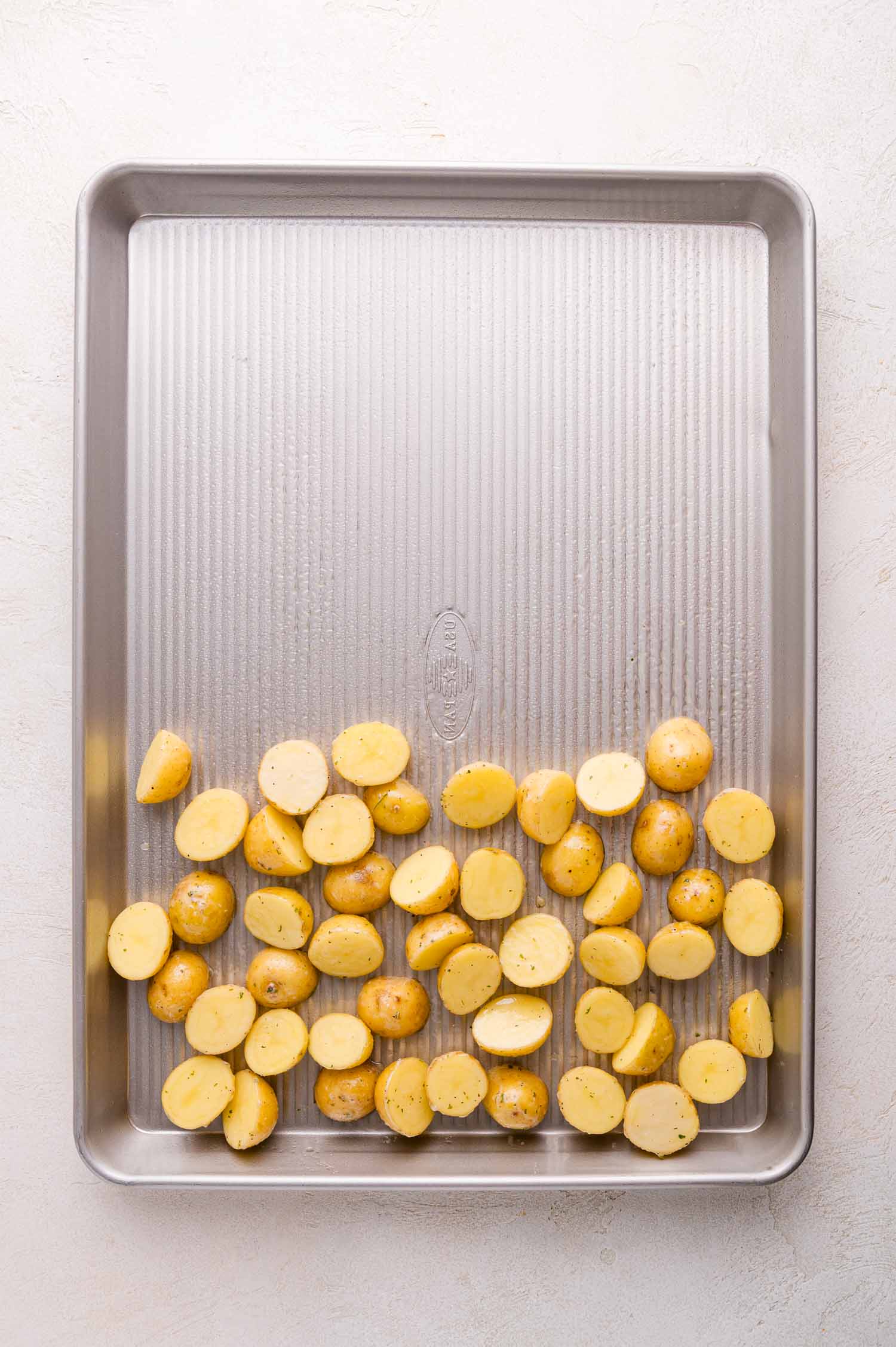 Halved baby potatoes spread in a single layer on a large, empty metal baking sheet.