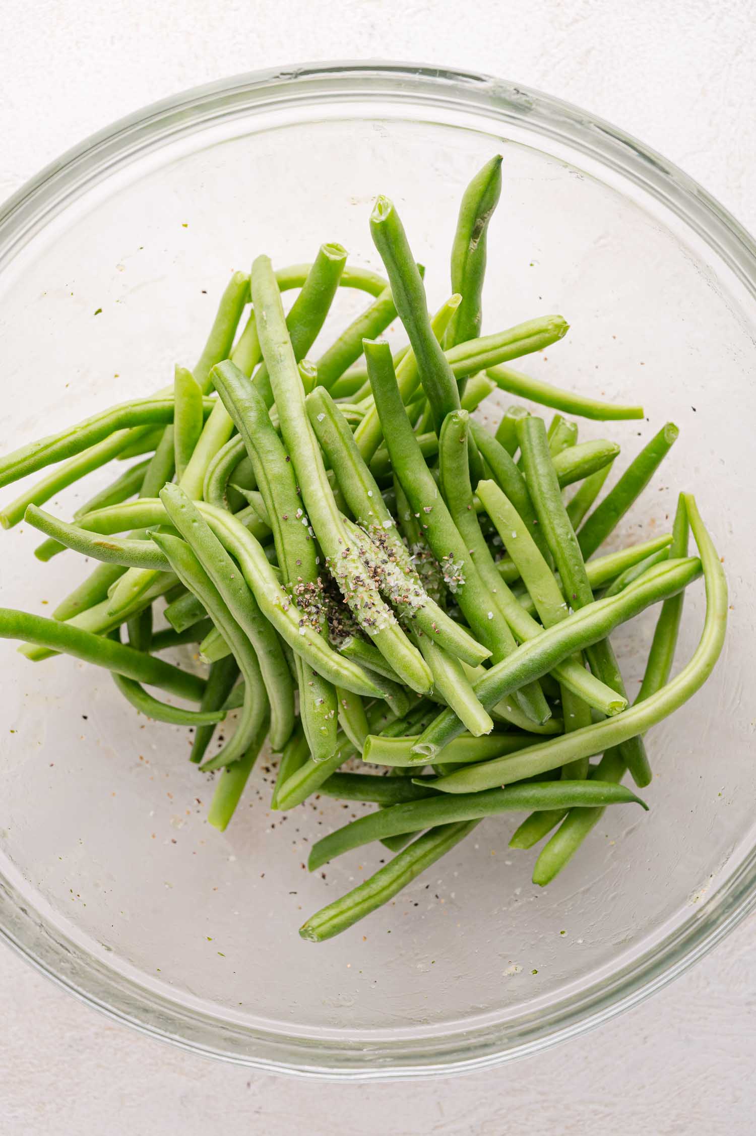 A glass bowl filled with fresh green beans, lightly sprinkled with salt and pepper.