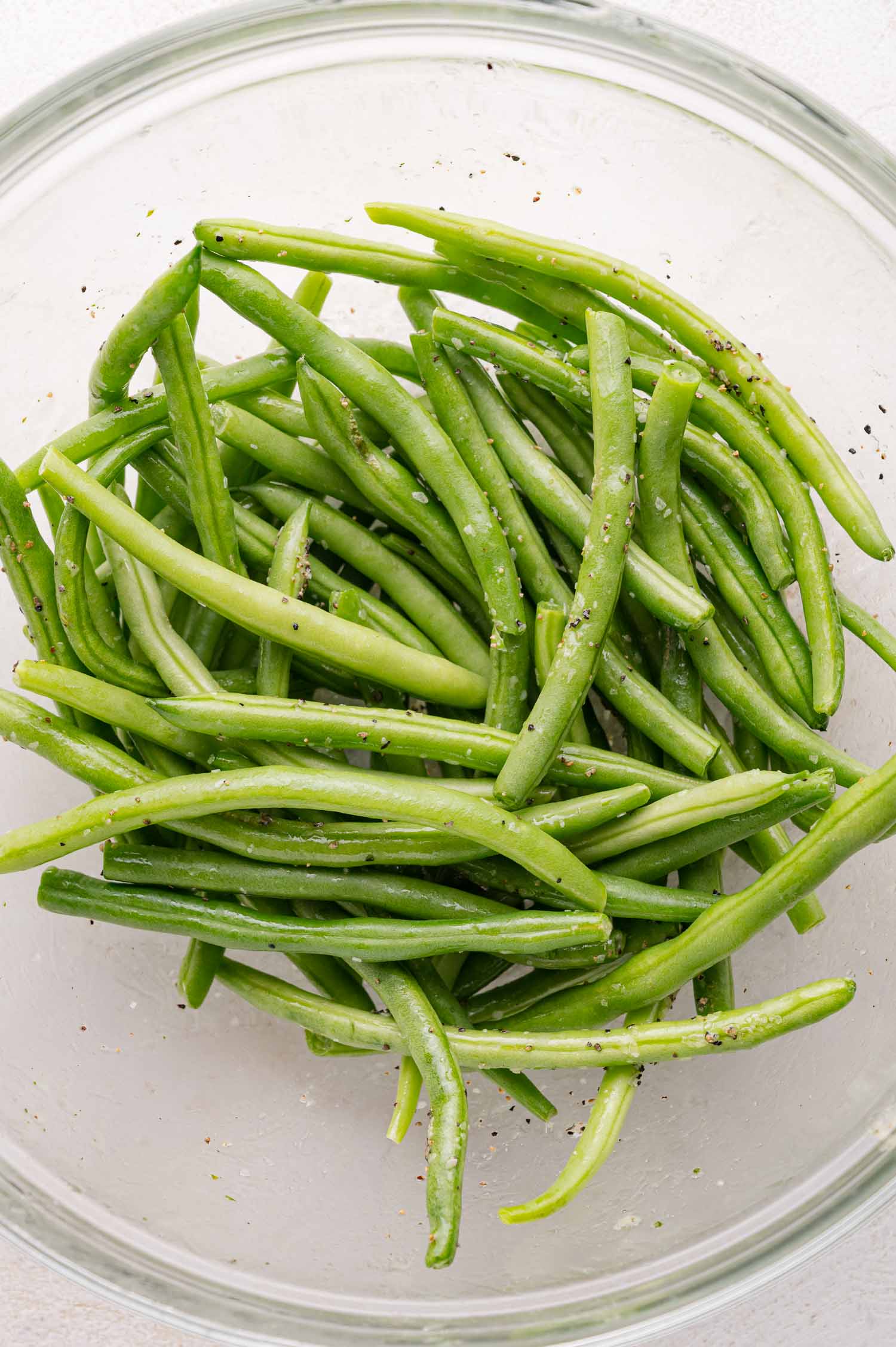 A clear glass bowl filled with fresh green beans seasoned with salt and pepper.