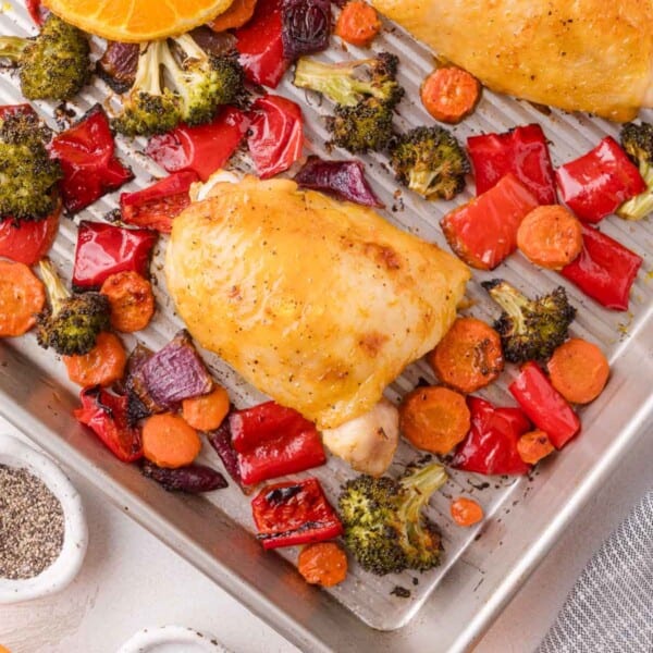 Baked chicken thighs with roasted red bell peppers, broccoli, carrots, and red onions on a metal baking tray.