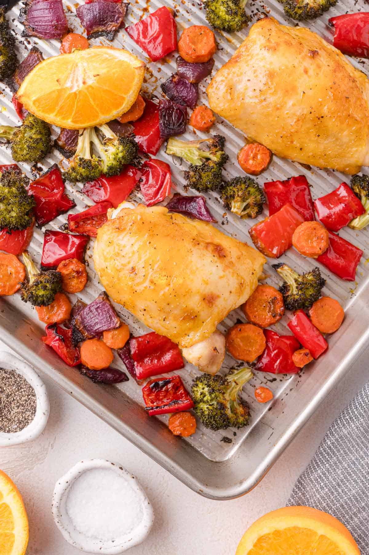 Baked chicken thighs with broccoli, carrots, red bell pepper, red onion, and orange slices on a sheet pan, seasoned with salt and pepper.