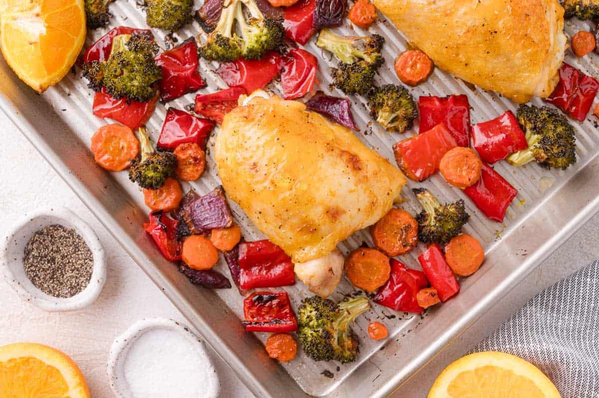 Baked chicken thighs with roasted red bell peppers, broccoli, carrots, and onions on a metal baking sheet, next to small bowls of salt and pepper and orange slices.