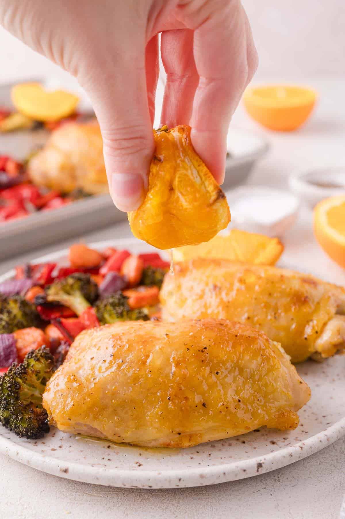 A hand squeezes an orange slice over roasted chicken thighs and vegetables on a plate, with more food and orange halves in the background.