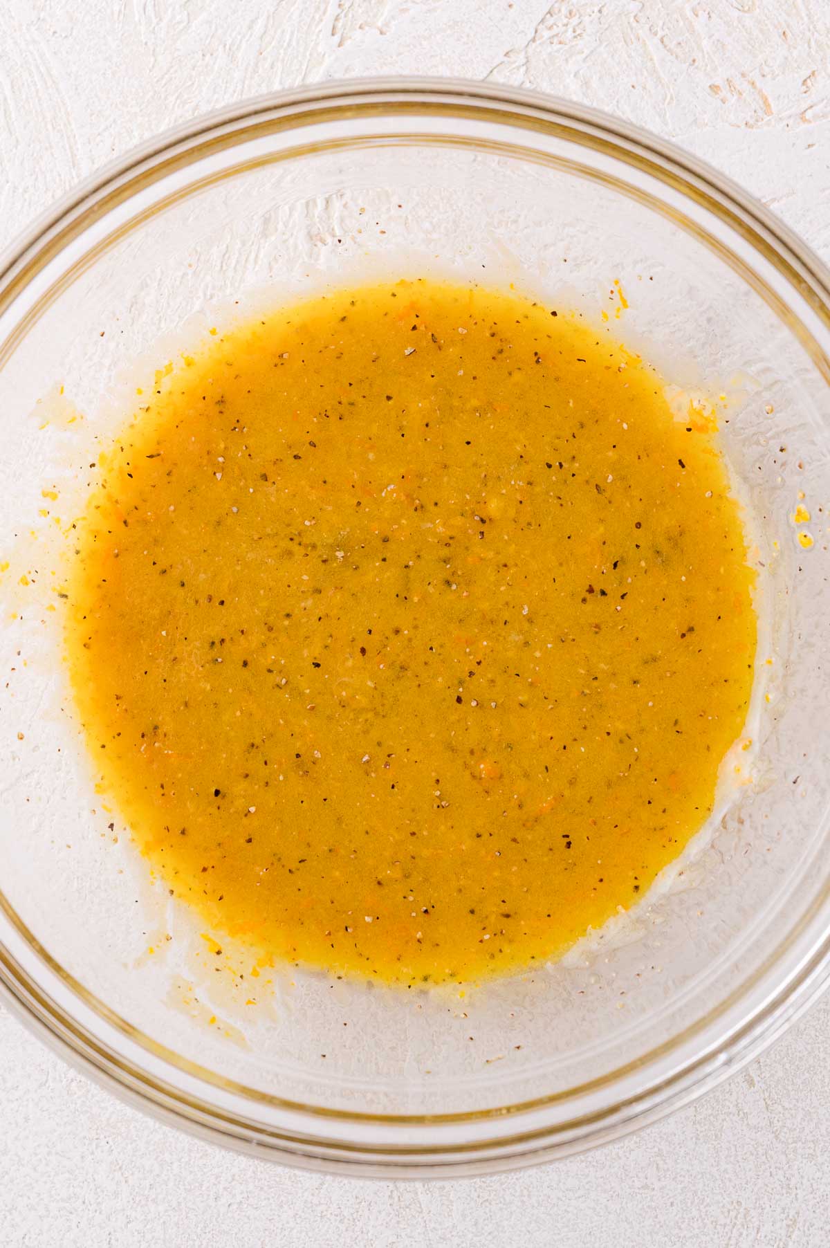 A glass bowl containing a yellow, speckled liquid mixture on a light textured surface.