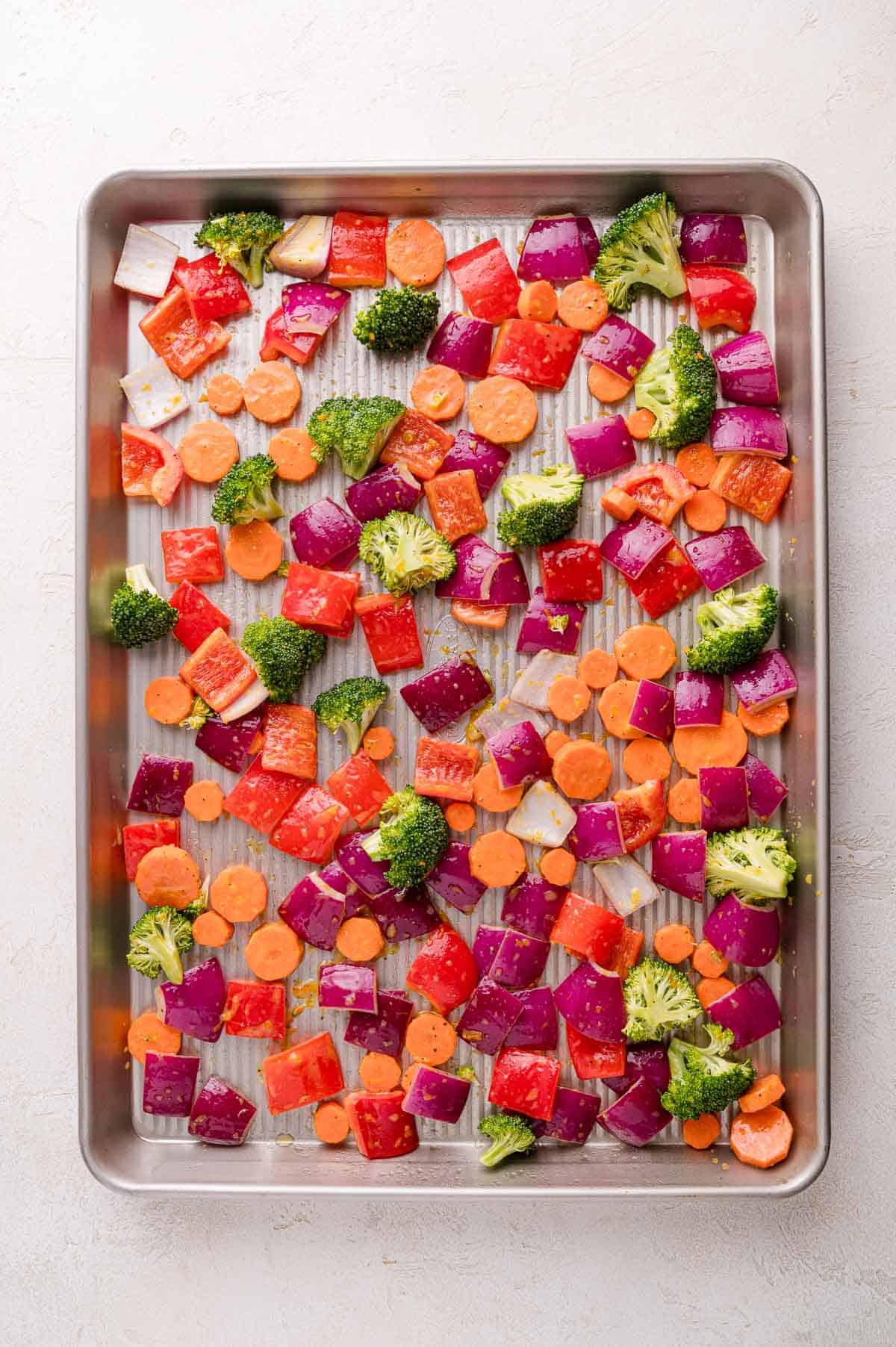 Chopped red onions, carrots, red bell peppers, and broccoli florets spread out on a metal baking sheet, ready for roasting.