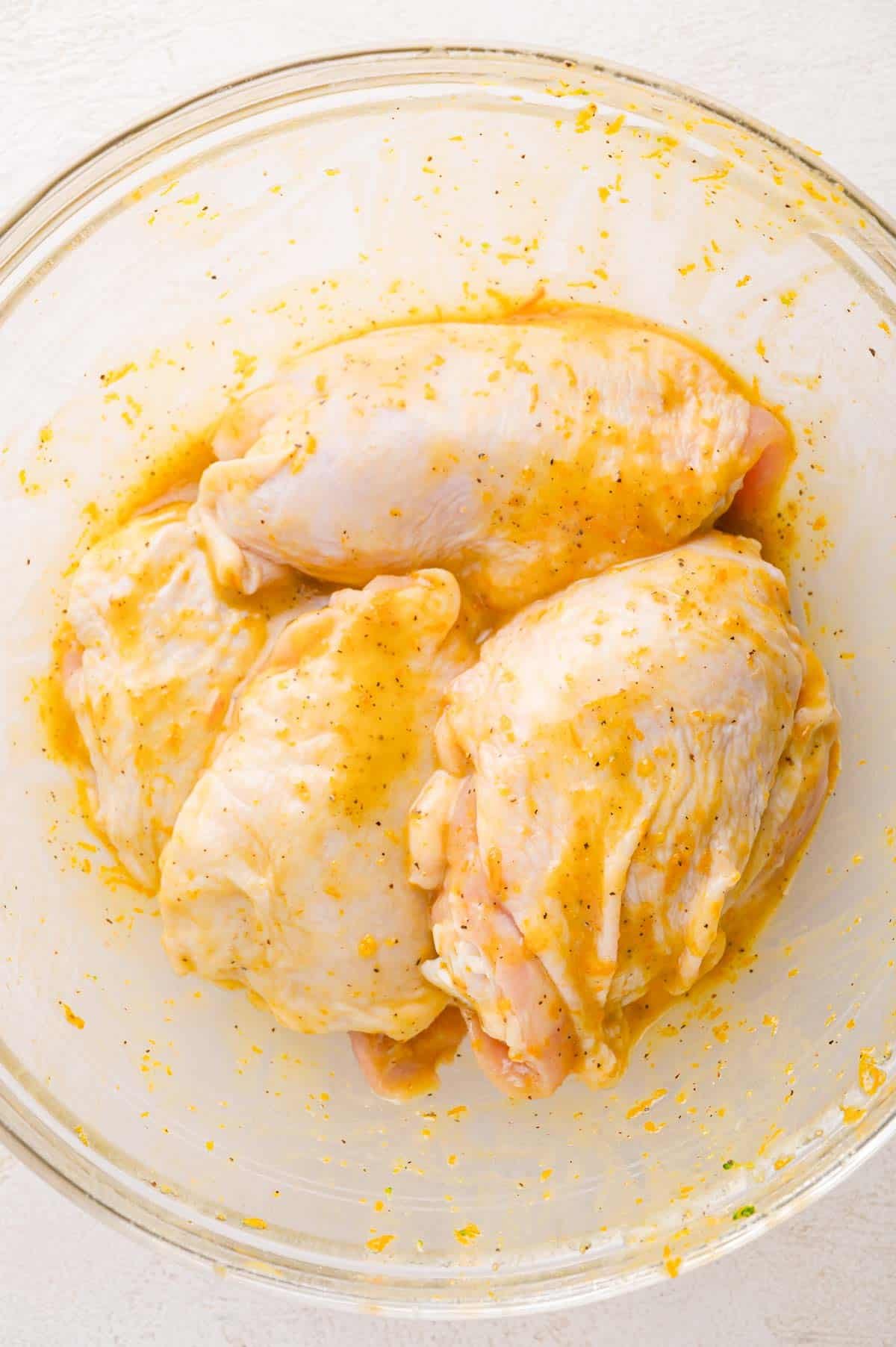 Raw chicken breasts coated in a yellow marinade with spices in a clear glass bowl on a white surface.