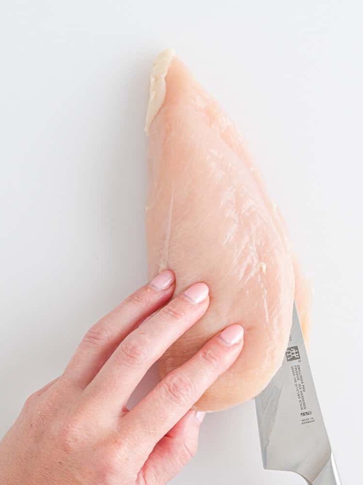 A hand holds a raw chicken breast on a white surface while slicing it horizontally with a kitchen knife.